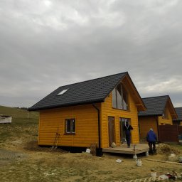 Usługi budowlane i ogrodnicze - Żółty dom z czarnym dachem i lukarną, widok z zewnątrz w pochmurny dzień. Częściowo wykończony taras i teren wokół domu.