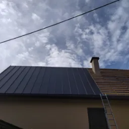 Częściowo pokryty dach blachą na rąbek stojący, widoczne łaty i komin na tle nieba z chmurami. Drabina oparta o ścianę budynku.