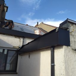 Usługi budowlane i ogrodnicze - Prace dekarskie na dachu budynku z widoczną nową, czarną obróbką blacharską i pracownikiem w trakcie montażu. Widoczne elementy konstrukcji dachu i niebo.