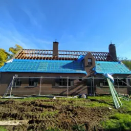 Dom z cegły w trakcie wymiany dachu: widoczne rusztowania, niebieska plandeka i drewniana konstrukcja dachu. Prace dekarskie w toku, słoneczny dzień.