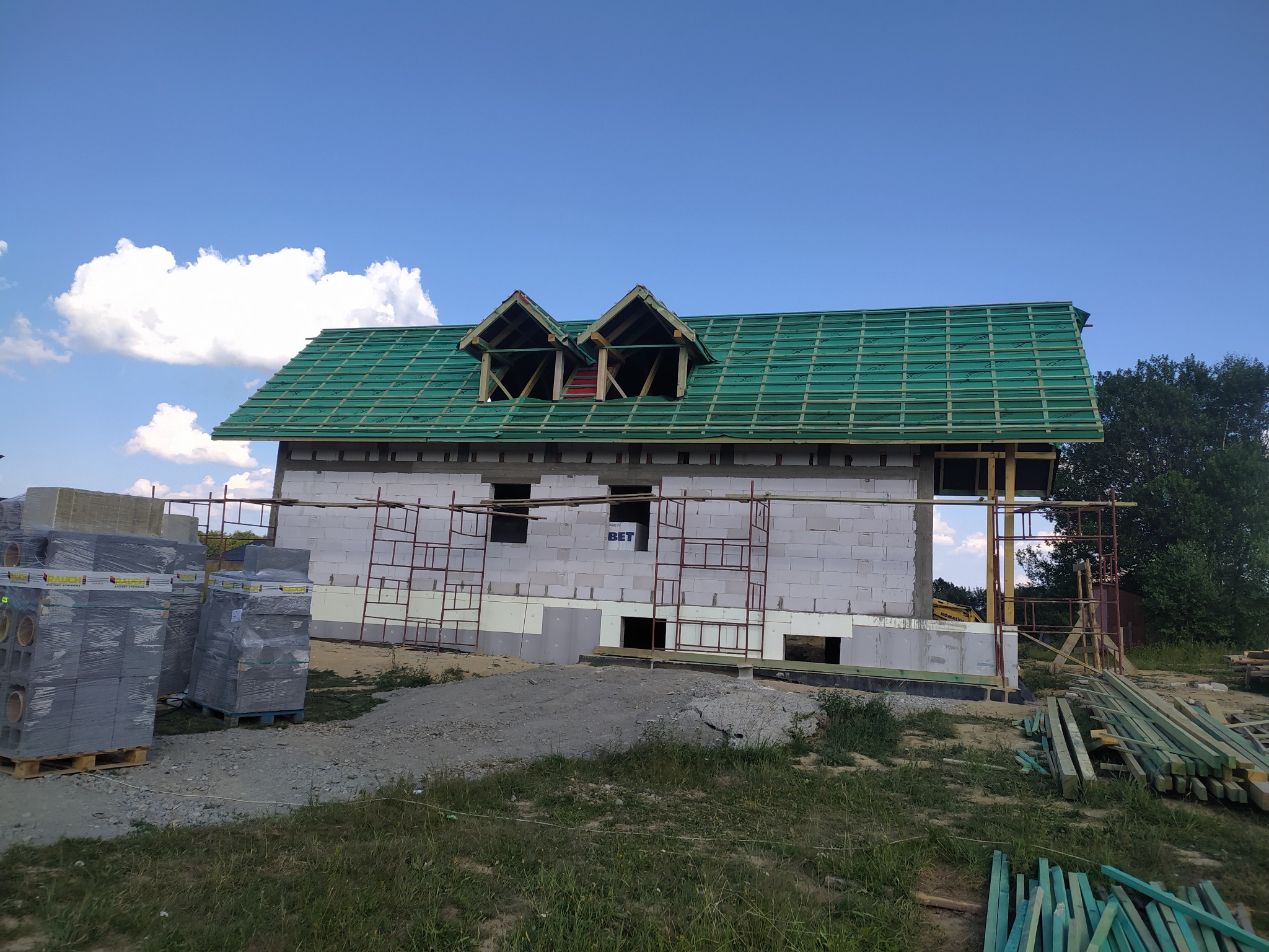 Dom w budowie z zielonym dachem w Jeleniej Górze. Widoczne materiały budowlane i rusztowania. Dachówka częściowo ułożona, okna dachowe w trakcie montażu.