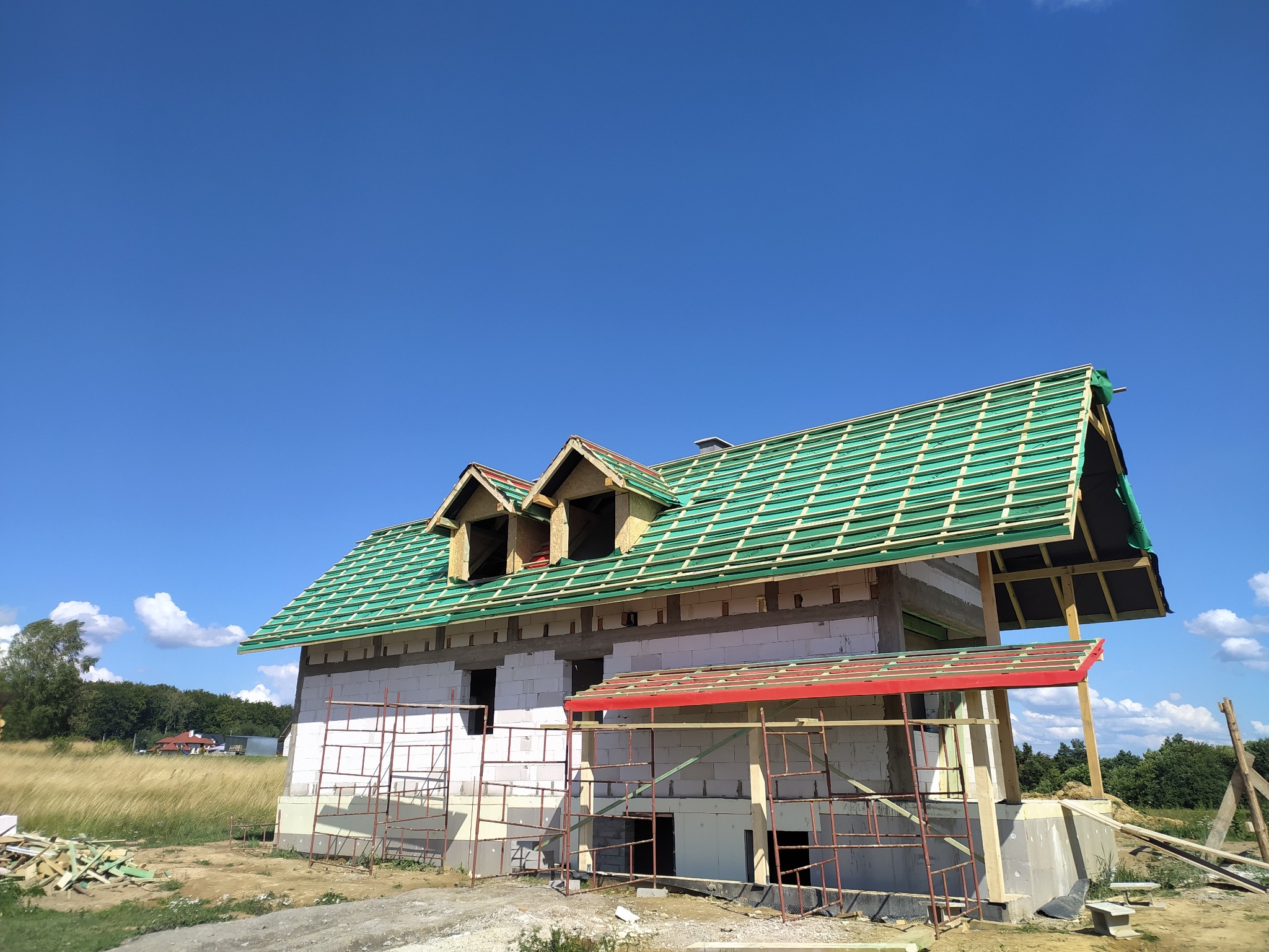 Niedokończony dom z zielonymi łatami na dachu i czerwonym zadaszeniem werandy, w słoneczny dzień. Widoczne rusztowania i materiały budowlane.