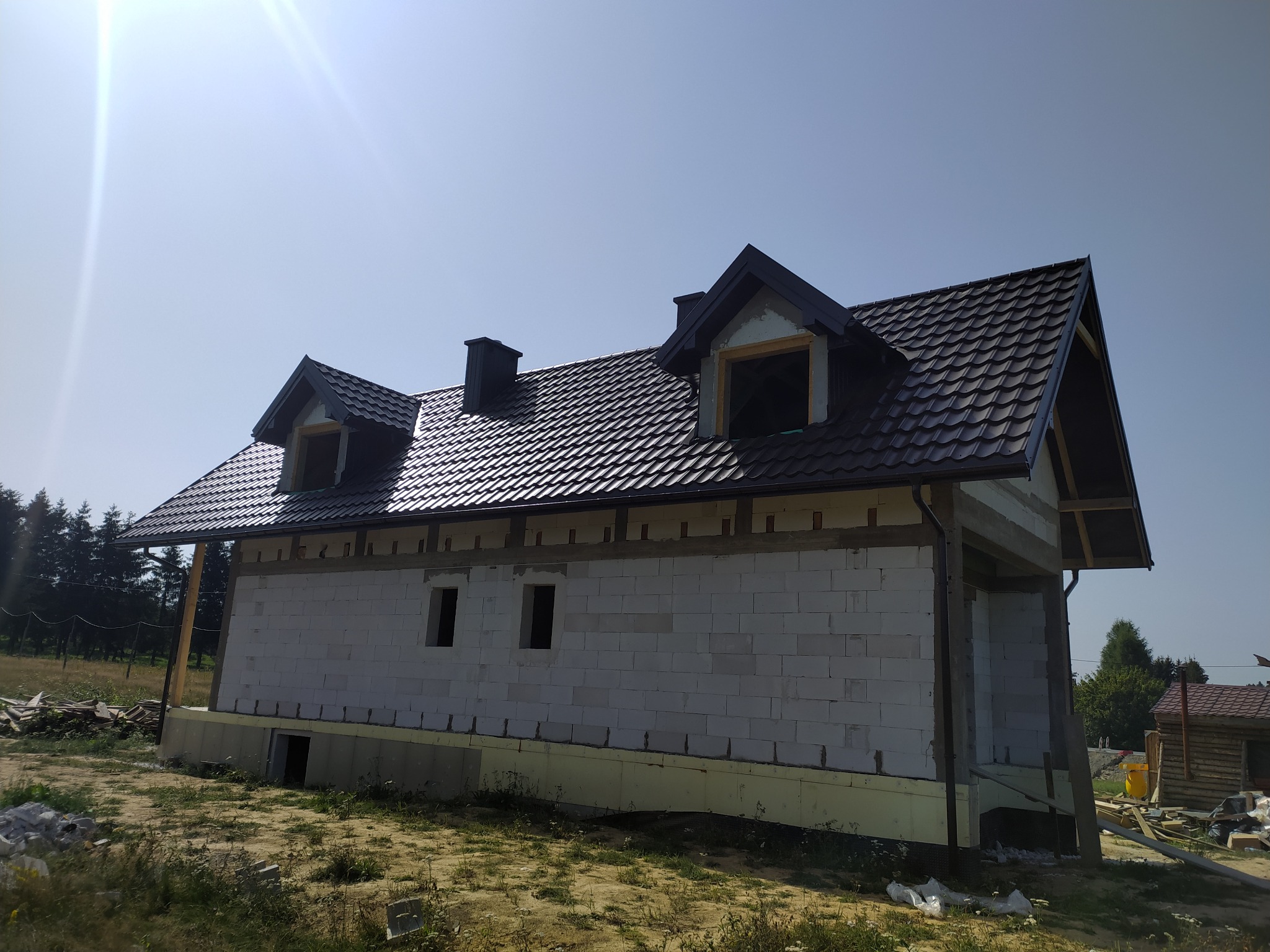 Dom w trakcie budowy z ciemnym, metalowym dachem i oknami dachowymi. Widoczne białe bloczki i elementy konstrukcyjne. Tło stanowi błękitne niebo i zieleń drzew.