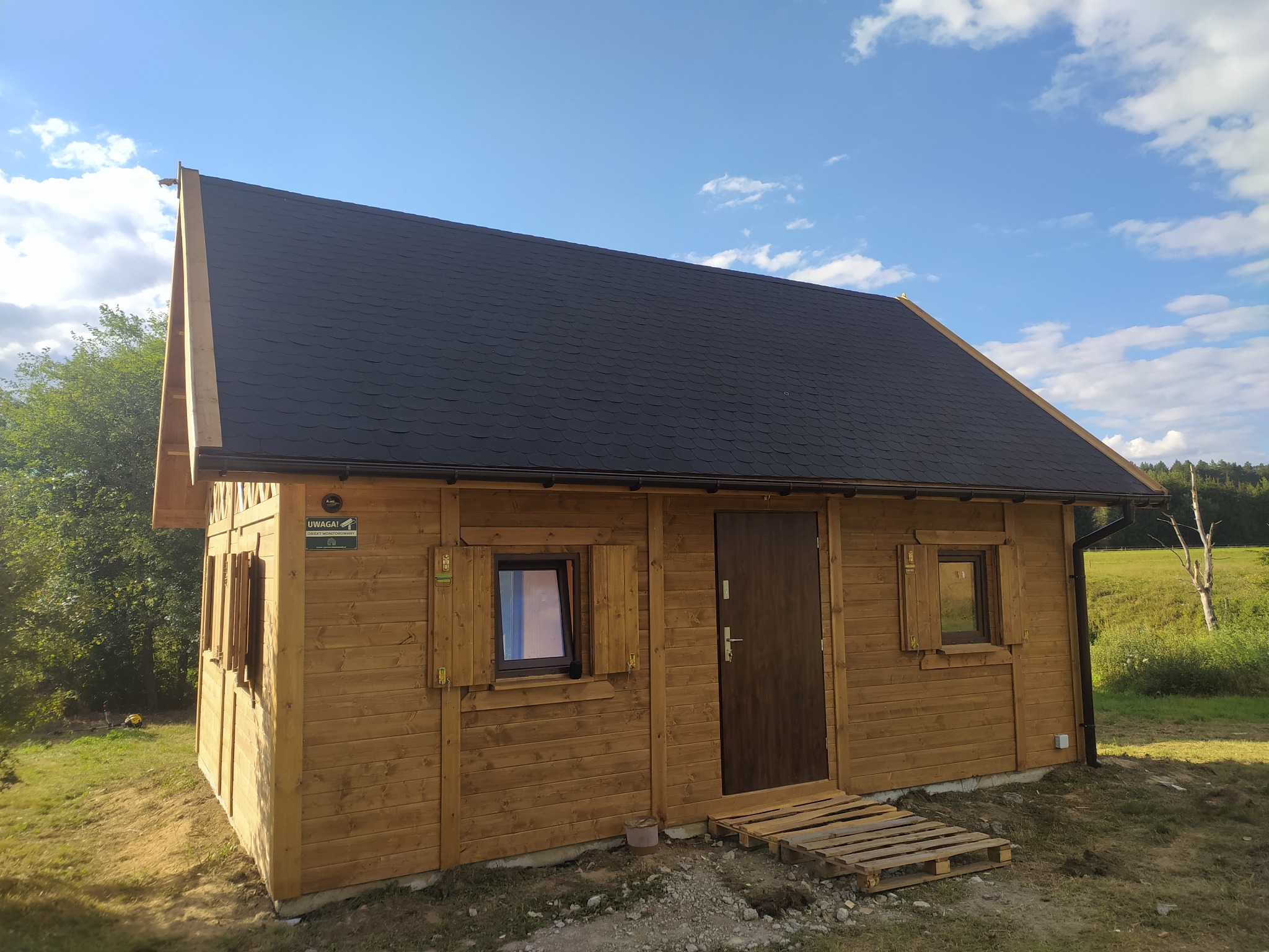 Drewniany domek z czarnym dachem z gontu bitumicznego, widok z zewnątrz, błękitne niebo w tle. Widoczne okna z okiennicami i paleta jako tymczasowe schody.