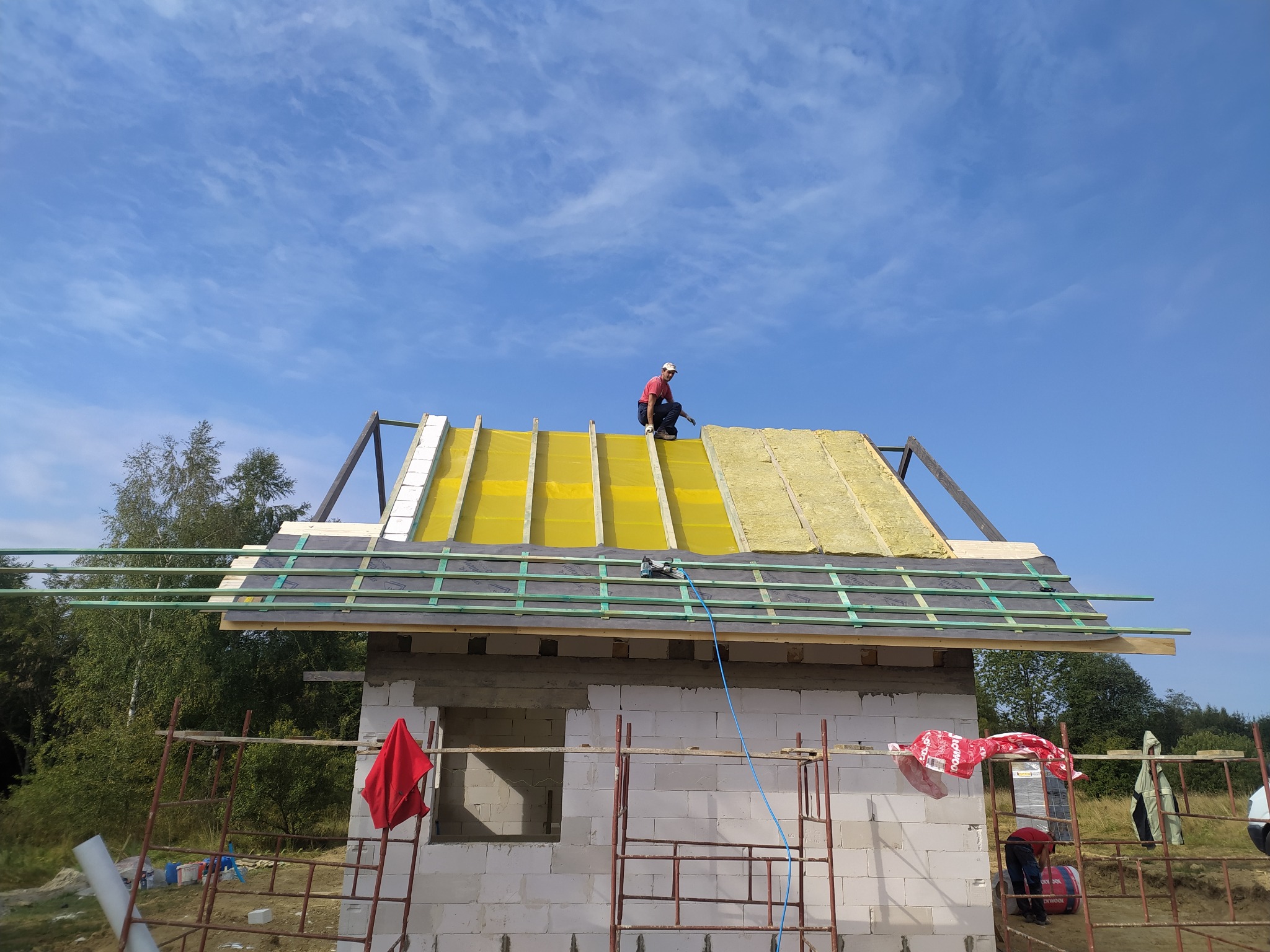 Montaż dachu na domu w budowie: dekarz na żółtej membranie i izolacji z wełny mineralnej, widoczne rusztowania i narzędzia dekarskie, błękitne niebo w tle.