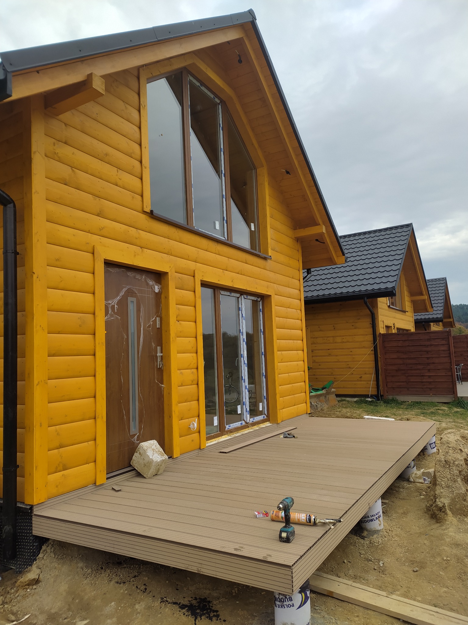 Żółty dom z drewnianą elewacją i czarnym dachem. Widoczny taras w trakcie budowy, narzędzia i materiały budowlane. Ujęcie z boku, w tle kolejne domy.
