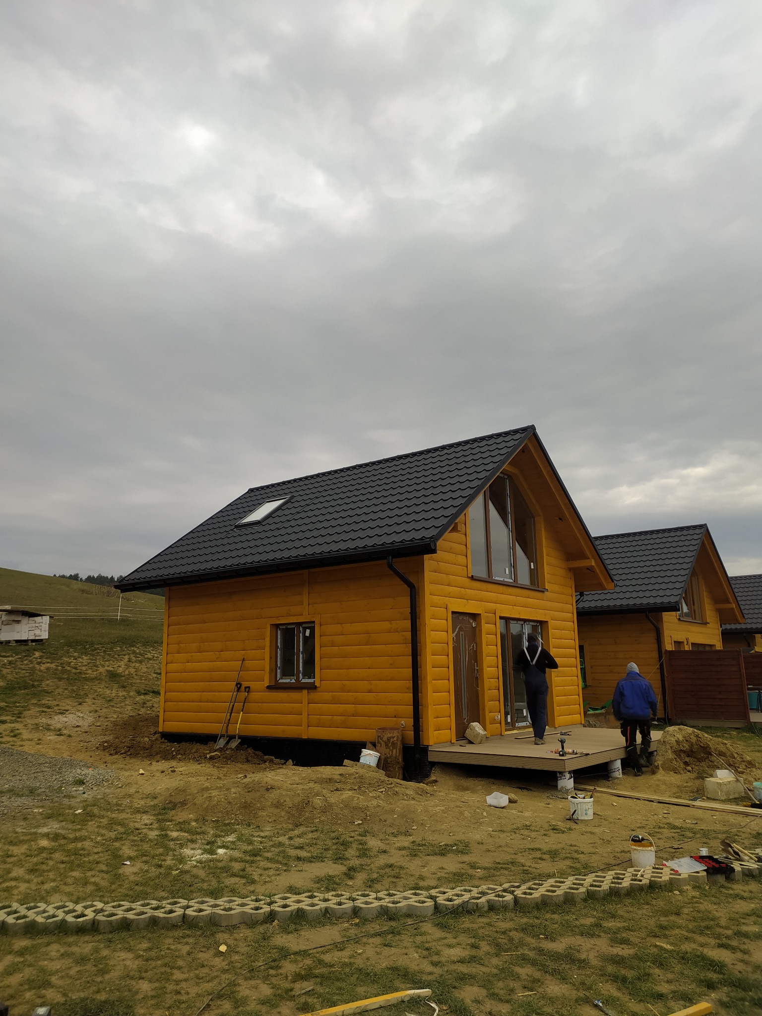 Żółty dom z czarnym dachem i lukarną, widok z zewnątrz w pochmurny dzień. Częściowo wykończony taras i teren wokół domu.