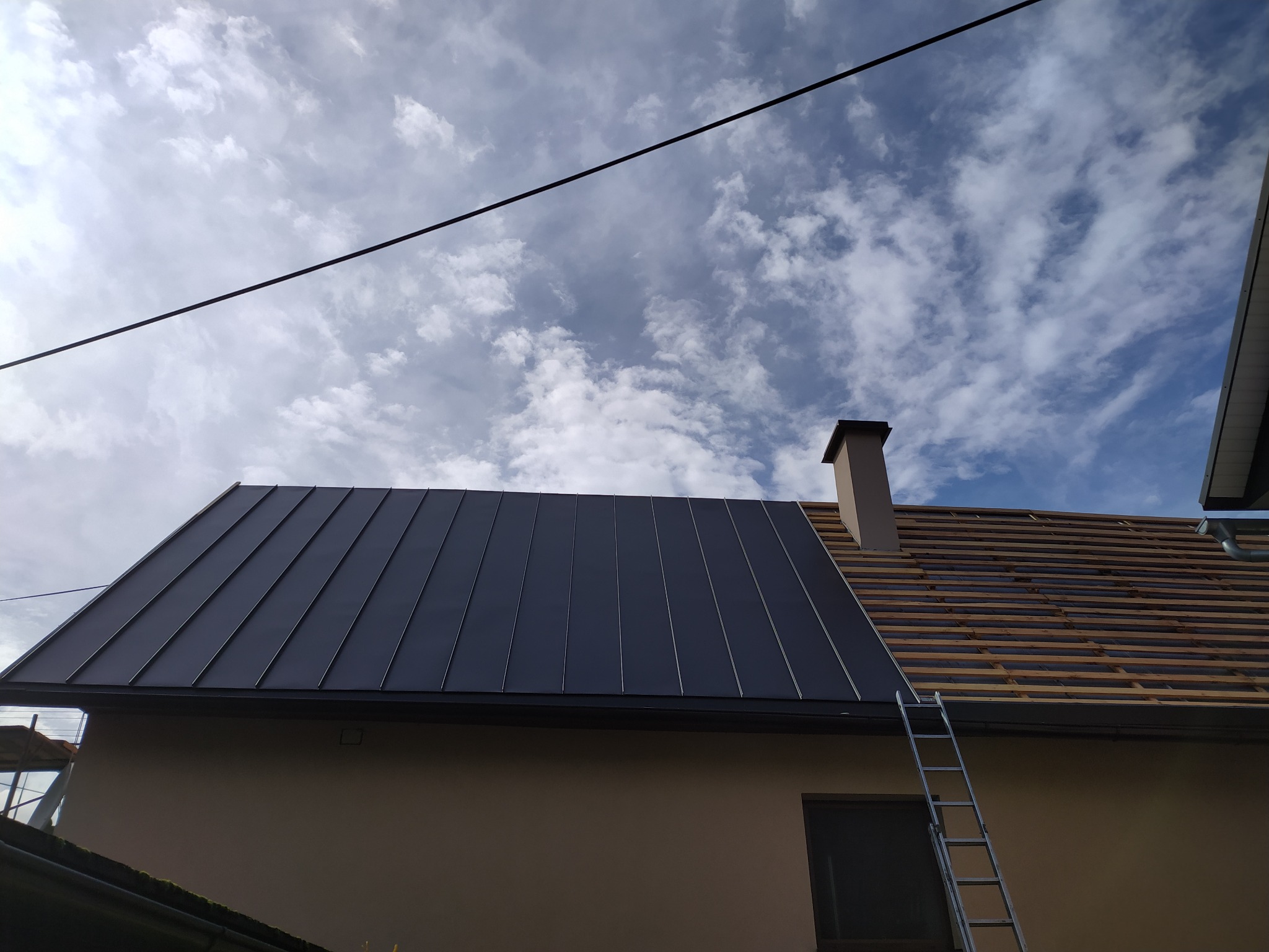 Częściowo pokryty dach blachą na rąbek stojący, widoczne łaty i komin na tle nieba z chmurami. Drabina oparta o ścianę budynku.