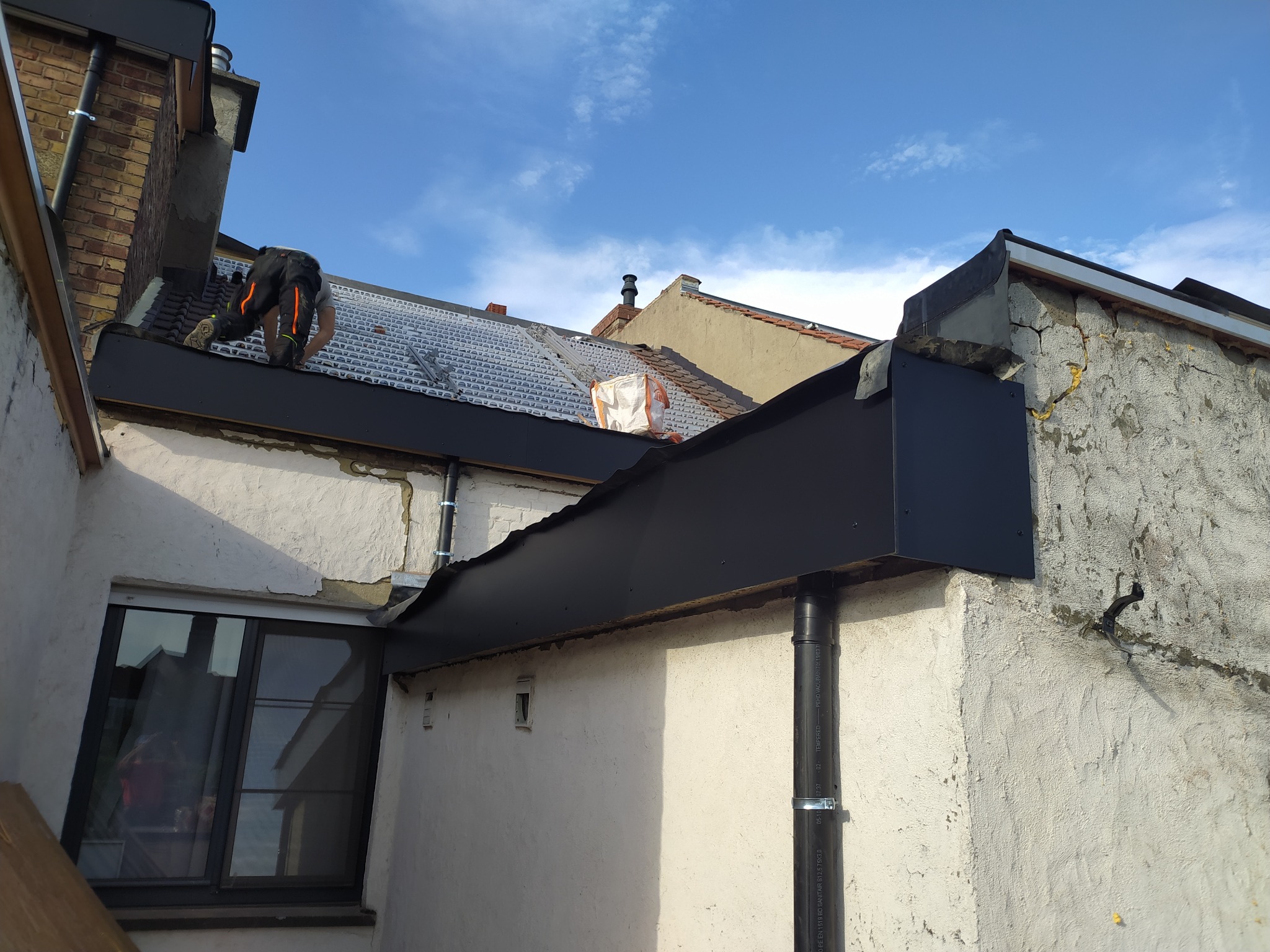 Prace dekarskie na dachu budynku z widoczną nową, czarną obróbką blacharską i pracownikiem w trakcie montażu. Widoczne elementy konstrukcji dachu i niebo.