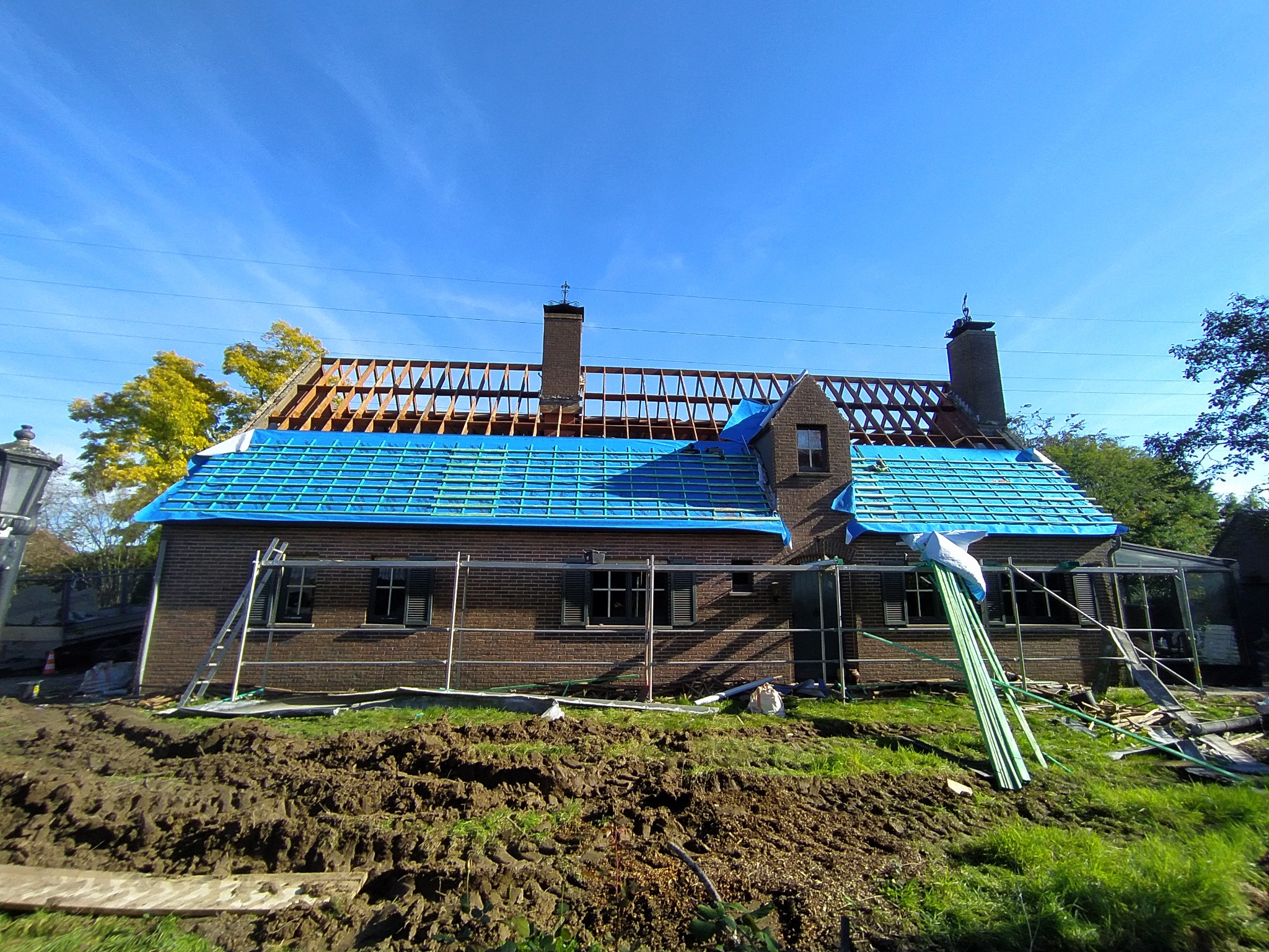 Dom z cegły w trakcie wymiany dachu: widoczne rusztowania, niebieska plandeka i drewniana konstrukcja dachu. Prace dekarskie w toku, słoneczny dzień.