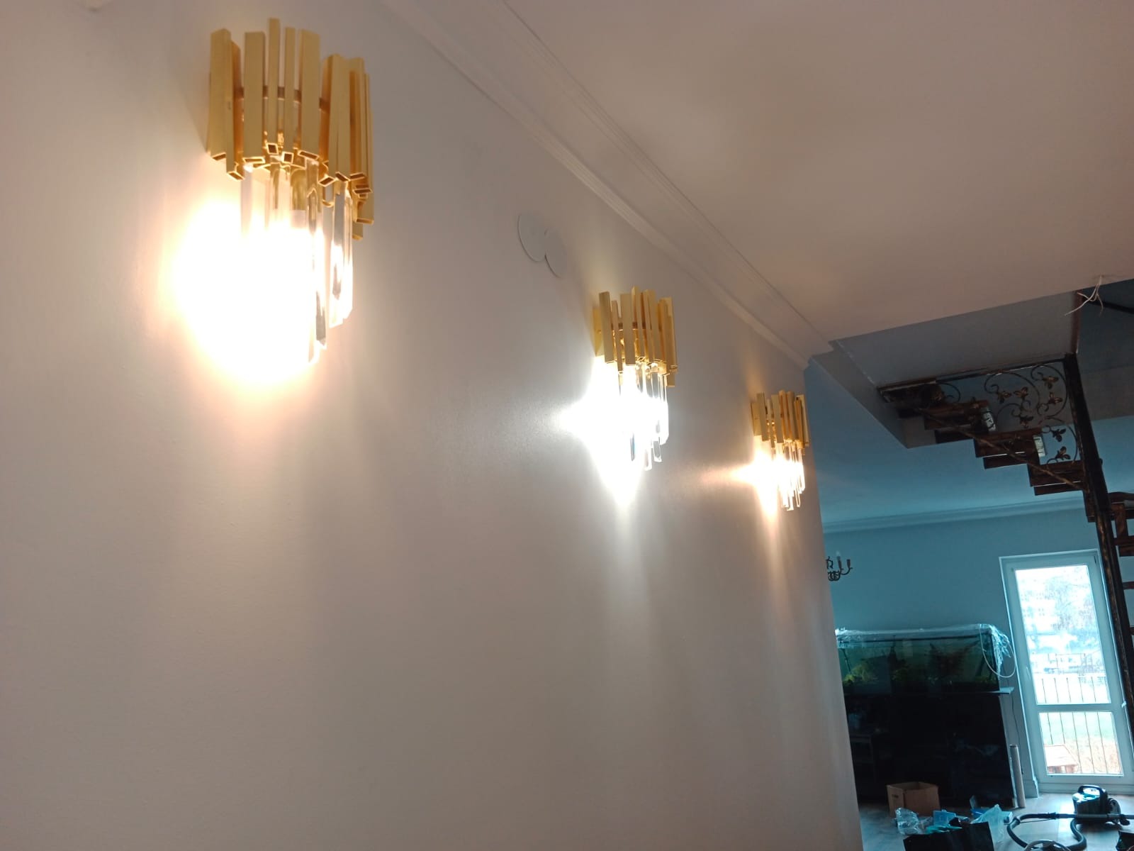 Świecące, złote kinkiety z akrylowymi elementami zamontowane na białej ścianie w rzędzie, oświetlające wnętrze z widocznymi schodami i akwarium.