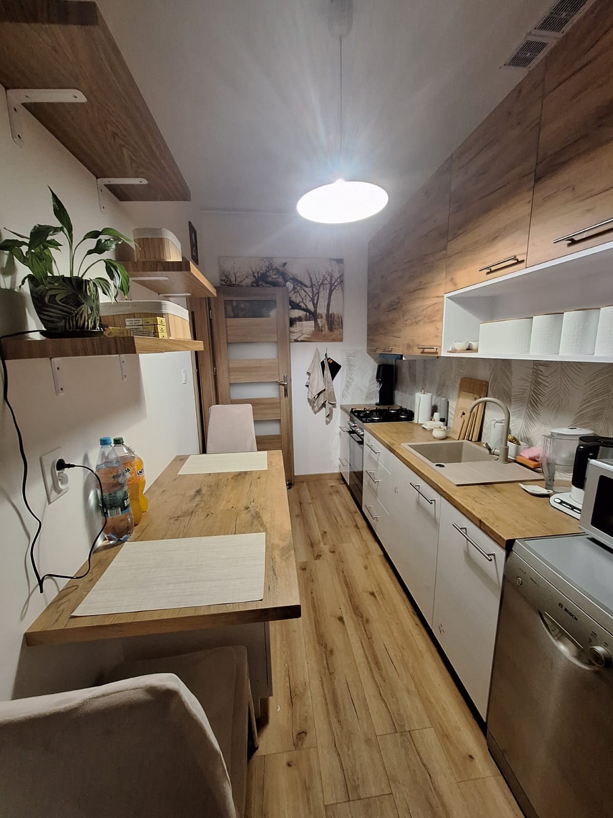 Nowoczesna kuchnia z drewnianymi akcentami: blat, podłoga, szafki górne w odcieniach drewna. Białe szafki dolne, stół z nakryciami, roślina doniczkowa i oświetlenie.