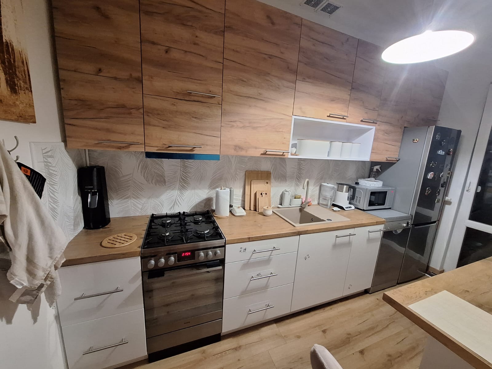 Nowoczesna kuchnia z drewnianymi szafkami górnymi i białymi dolnymi, z widoczną kuchenką gazową i elementami wyposażenia.