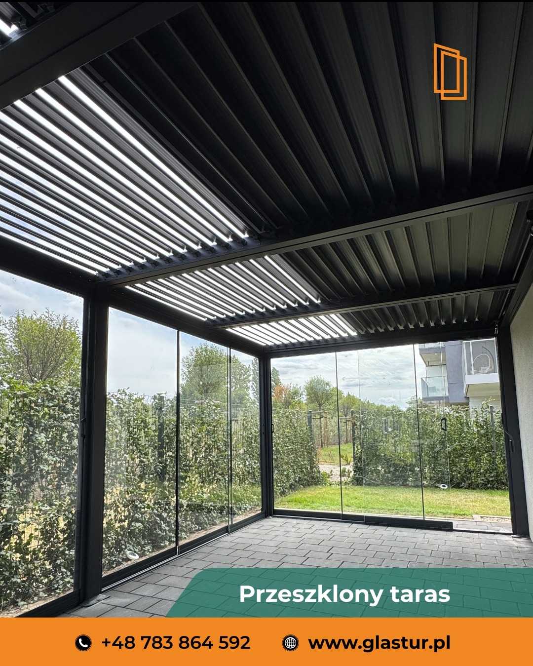 Przeszklony taras z metalową pergolą w kolorze grafitu. Widoczne szklane ściany przesuwne i żaluzjowy dach. Zewnętrzny widok na zieleń i budynki mieszkalne.