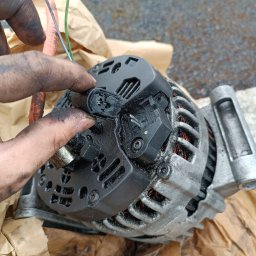 Kamil - Brudne ręce mechanika podłączają przewody do alternatora samochodowego na tle papieru pakowego. Widoczne detale konstrukcji alternatora i złącza.