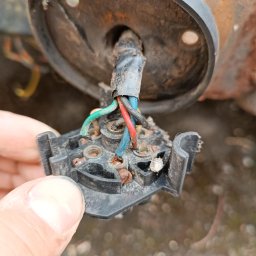 Kamil - Uszkodzona, skorodowana instalacja elektryczna z widocznymi przewodami w kolorach: zielonym, czerwonym i niebieskim, trzymana w dłoni. Widoczne ślady rdzy.