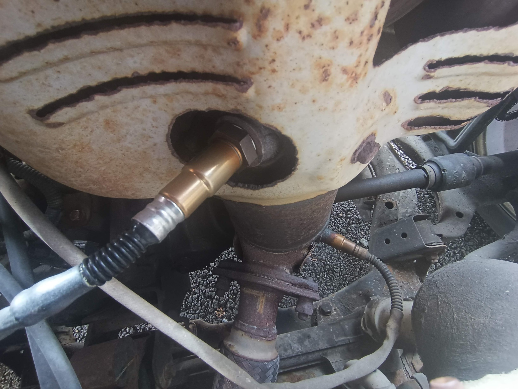 Zardzewiały układ wydechowy samochodu z widocznym czujnikiem tlenu i elementami podwozia na żwirowym podłożu. Detal z bliska, rdza, przewody.