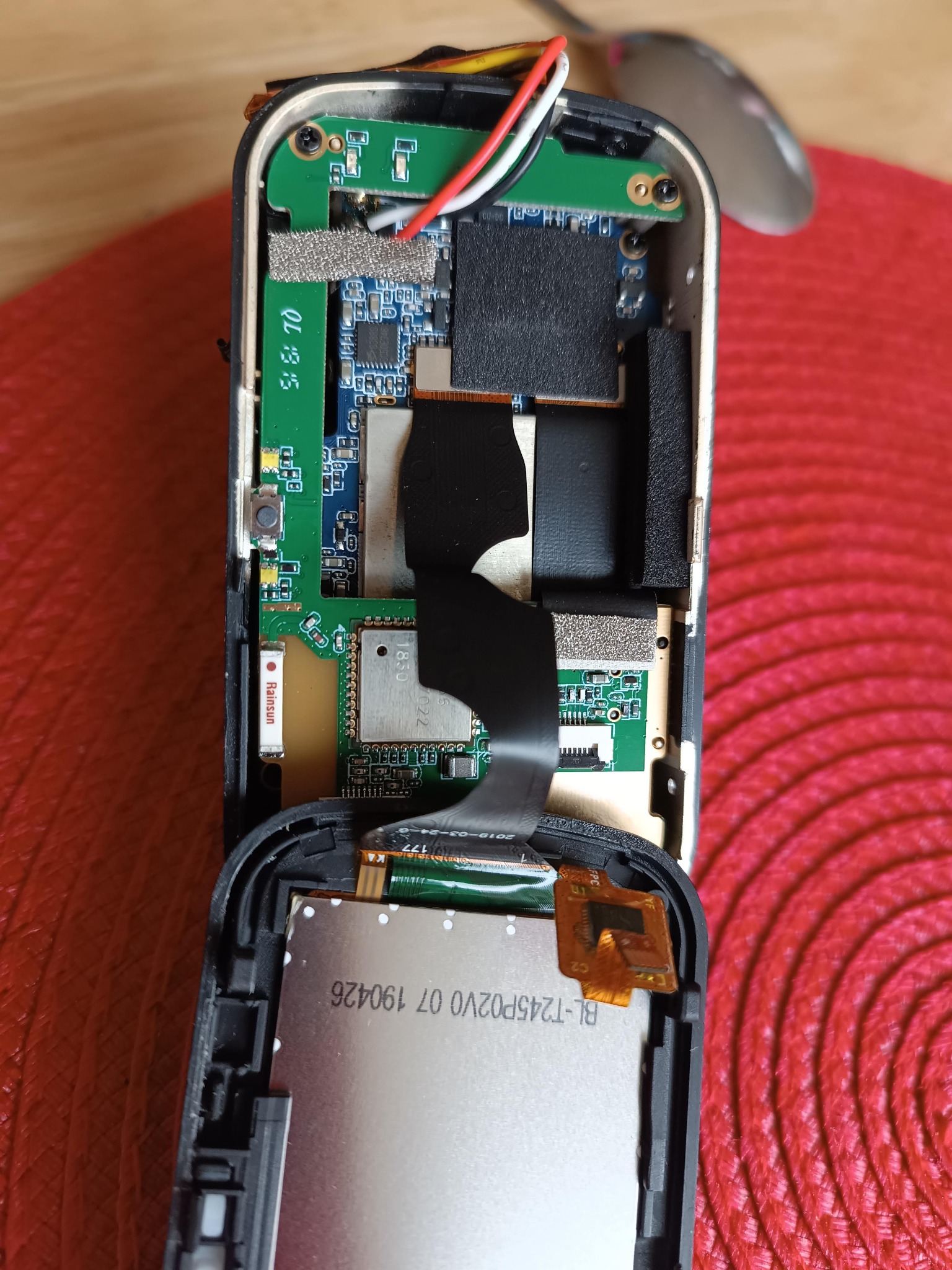 Otwarty smartfon z widoczną elektroniką wewnętrzną: płyta główna, układy scalone, taśmy połączeniowe. Widoczny ekran i fragment obudowy. Czerwone tło.