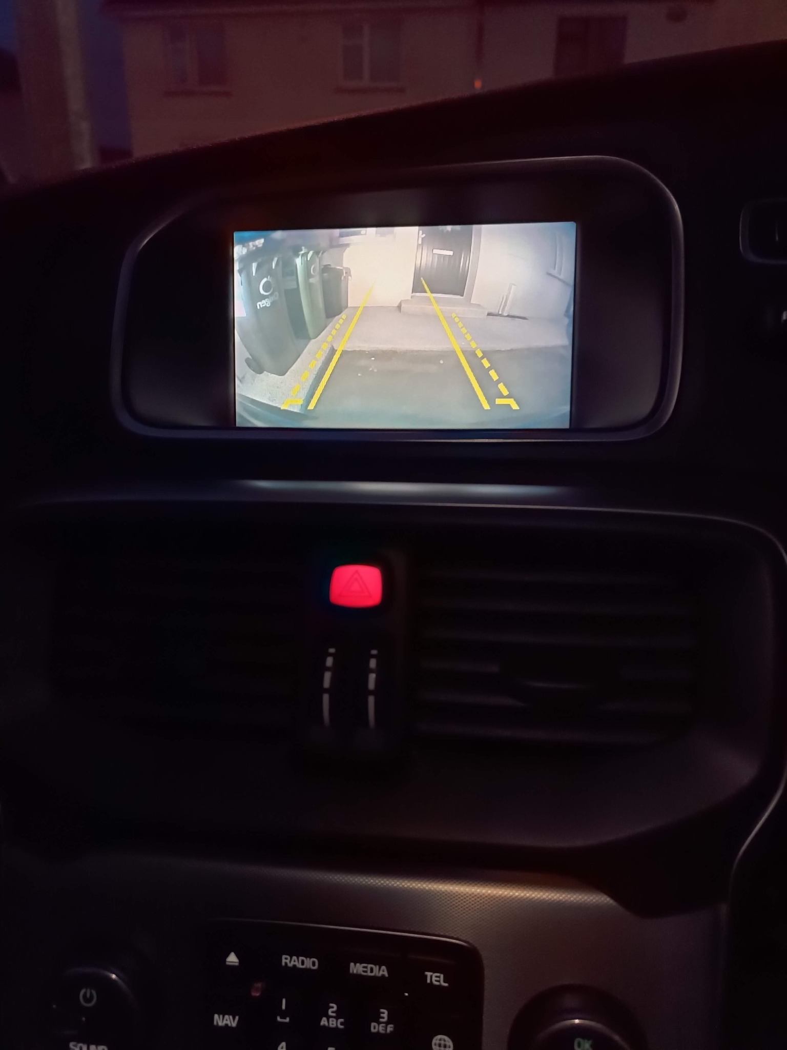 Wnętrze samochodu z widokiem na ekran kamery cofania, prezentujący obraz otoczenia z tyłu pojazdu. Widoczne linie pomocnicze ułatwiające parkowanie.