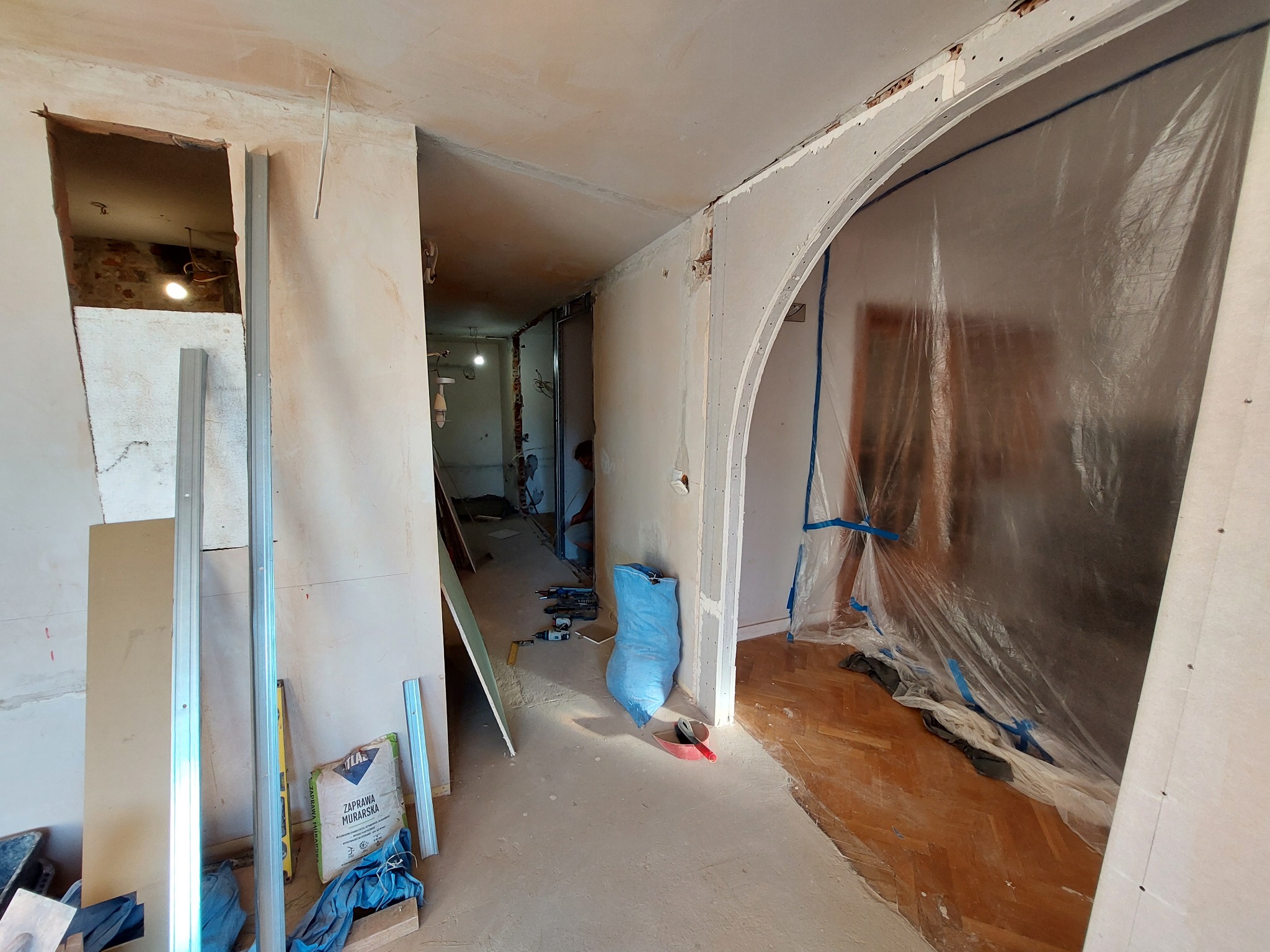 Wnętrze w trakcie remontu: postawiona ścianka działowa z płyty g-k z wyciętym łukiem, zabezpieczona folią, widoczne profile, zaprawa murarska i narzędzia.