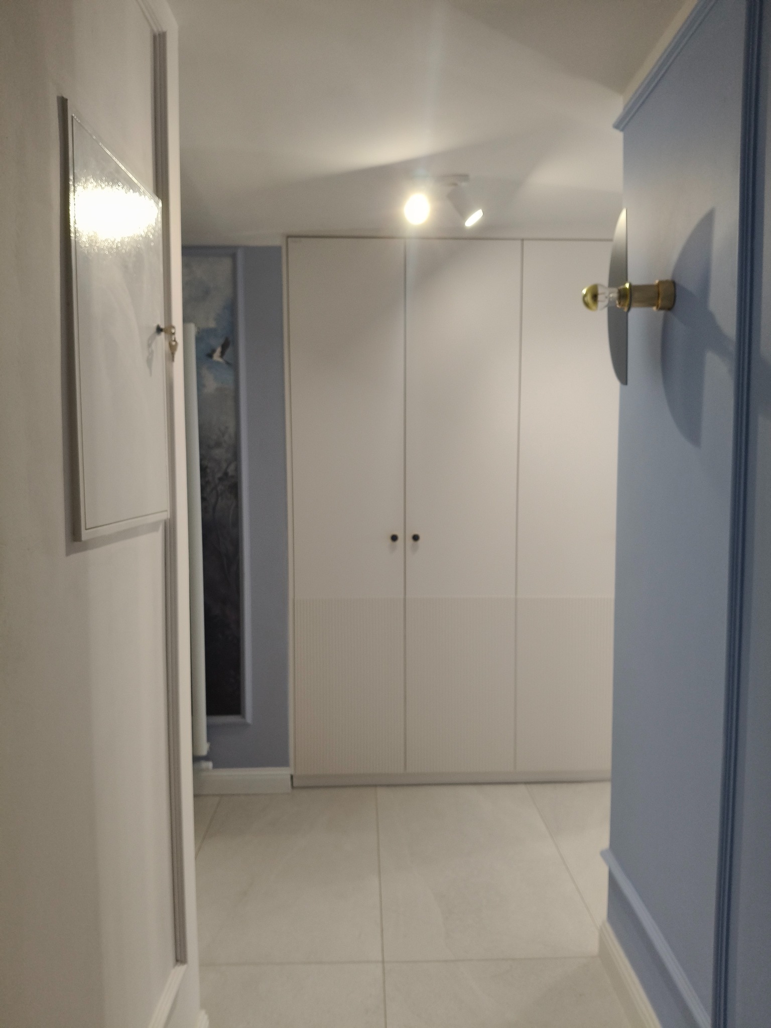 Nowoczesny przedpokój z białymi szafami w zabudowie, szarą podłogą i dekoracyjną lampą ścienną. Minimalistyczny design.