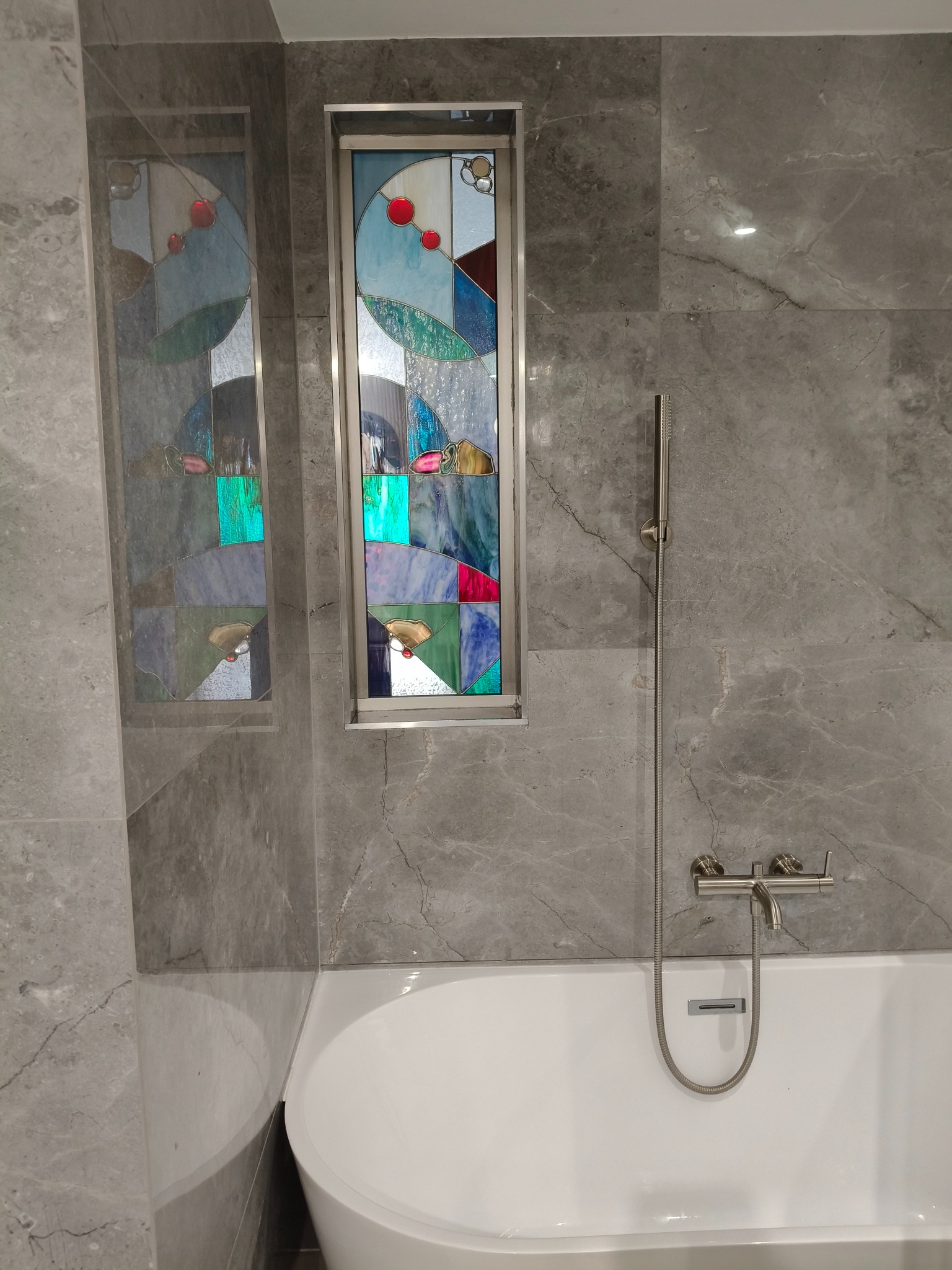 Szara łazienka z wanną i dekoracyjnymi witrażami w ścianie, podkreślającymi unikalny charakter wnętrza. Nowoczesna armatura.