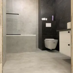 Minimalistyczna łazienka po remoncie: szare płytki imitujące beton, podwieszana toaleta i biała szafka z czarnymi uchwytami. Widoczne niedokończone obramowanie drzwi.