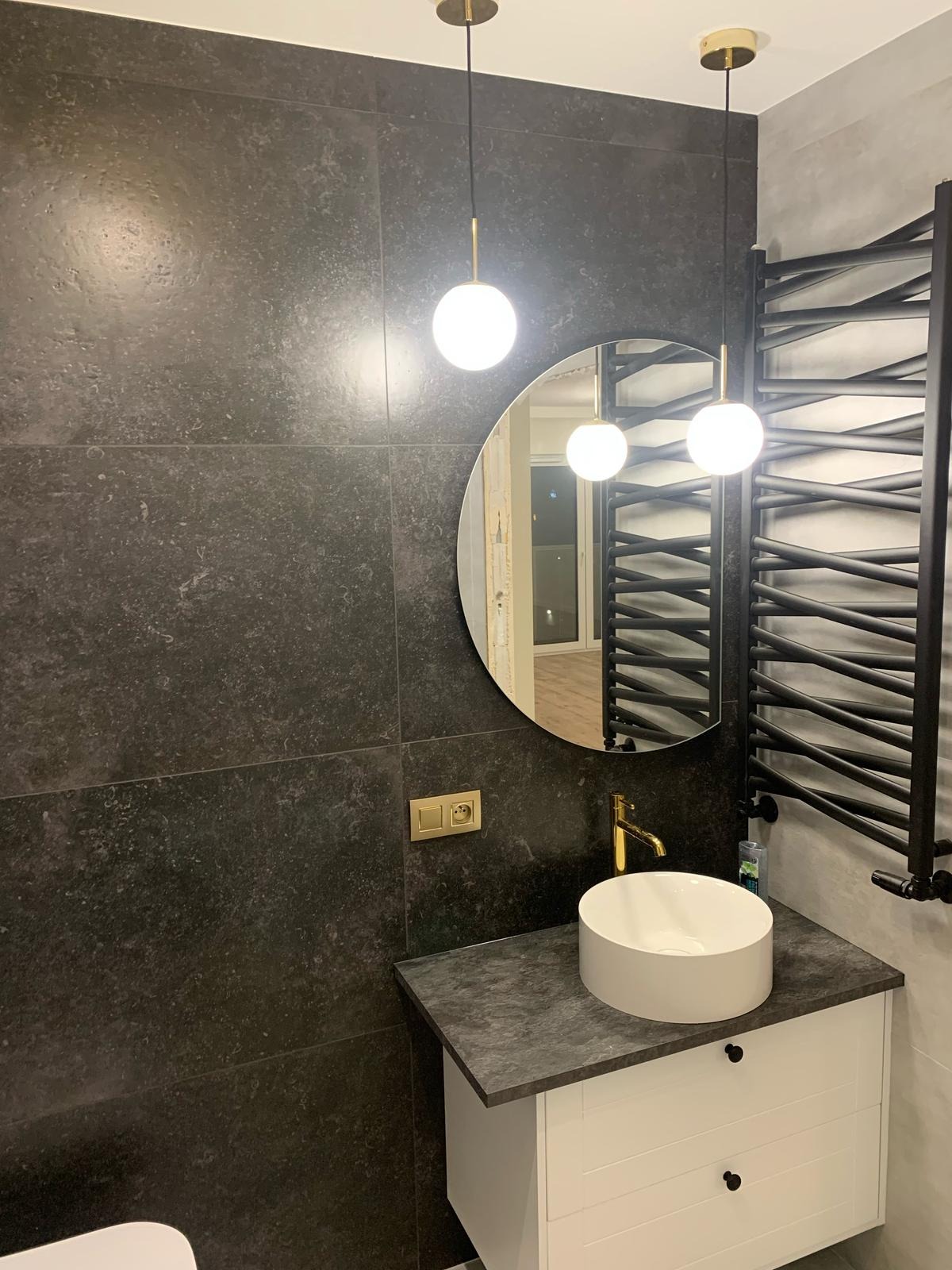 Nowoczesna łazienka z czarnymi płytkami, okrągłym lustrem, białą umywalką nablatową i grzejnikiem drabinkowym. Dwie wiszące lampy kuliste. Złota armatura.