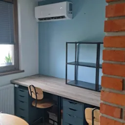 Klimatyzator ścienny zamontowany nad biurkiem z drewnianym blatem, obok metalowej półki na tle błękitnej ściany. Widoczny fragment ceglanej ściany.
