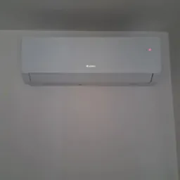 Biała klimatyzacja Gree, zamontowana na ścianie, z czerwoną diodą sygnalizacyjną. Minimalistyczny design, neutralne tło ściany.