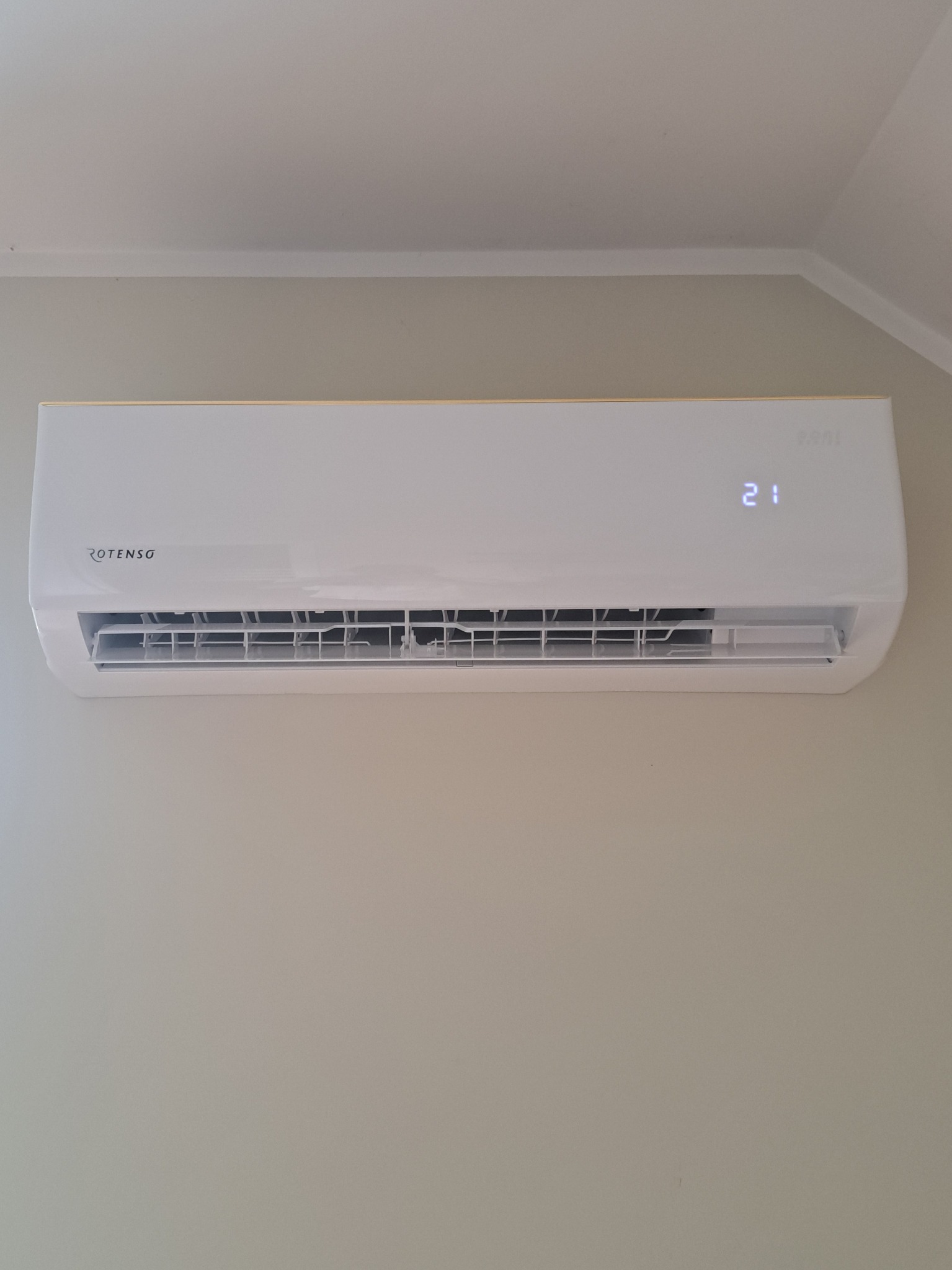 Biała klimatyzacja Rotenso zamontowana na ścianie, wyświetlająca temperaturę 21 stopni, widok z przodu, oświetlenie dzienne.