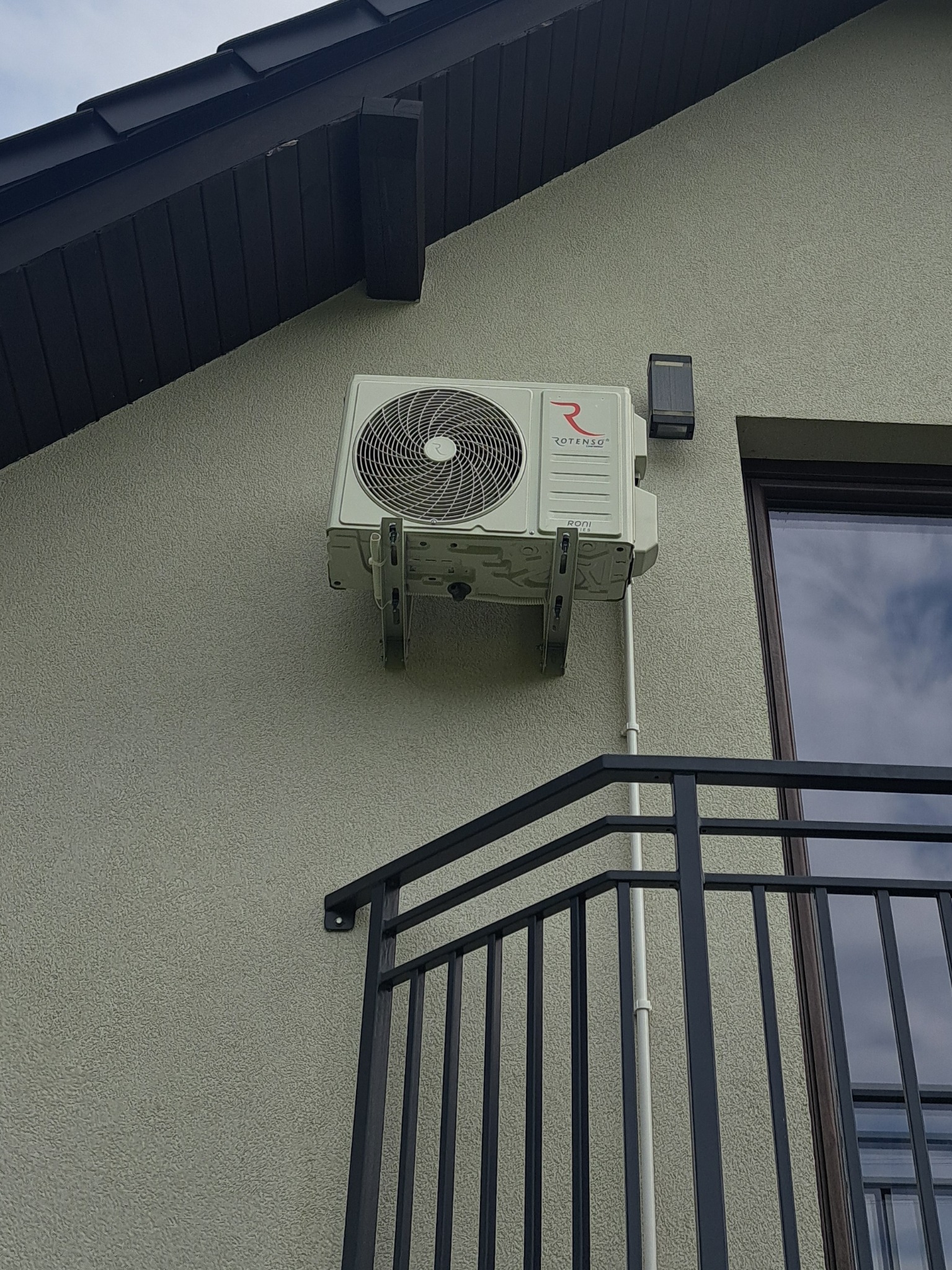 Zamontowana klimatyzacja Rotenso na elewacji domu, widoczna z perspektywy balkonu. Biała jednostka zewnętrzna z widocznym logo, rurami i elementami mocującymi.