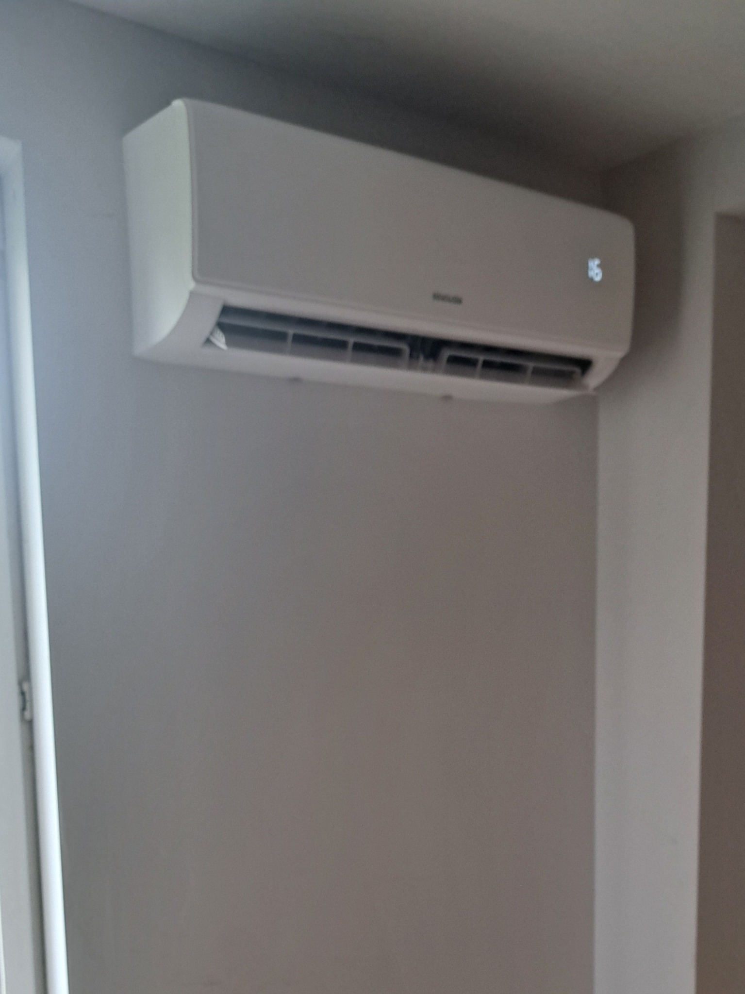 Biała klimatyzacja ścienna z wyświetlaczem temperatury, zamontowana na szarej ścianie w rogu pomieszczenia, widok z dołu.
