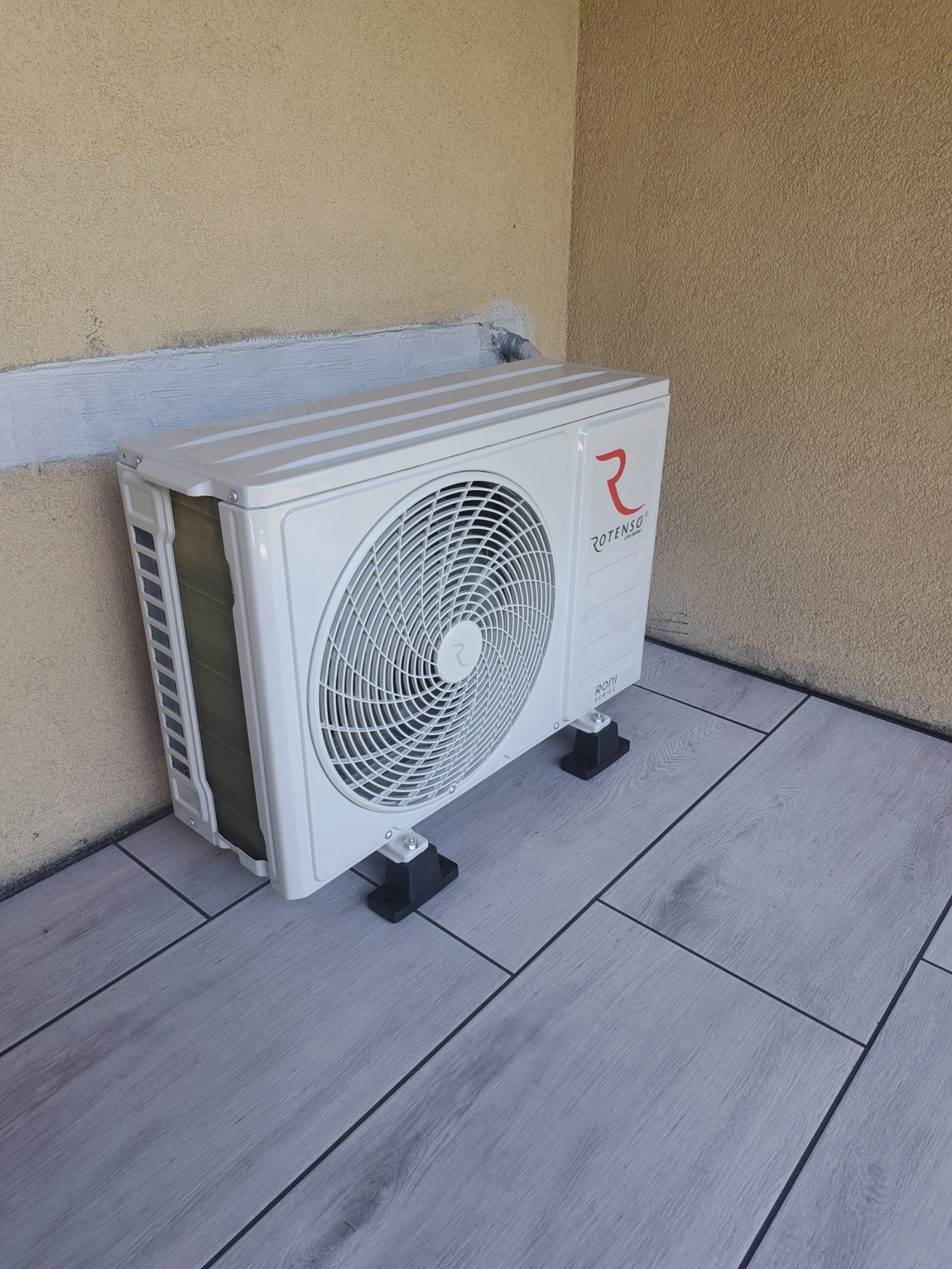 Biała jednostka zewnętrzna klimatyzacji Rotenso, model Rodi Series, zamontowana na balkonie z szarymi płytkami, przy beżowej ścianie. Widok z dołu.