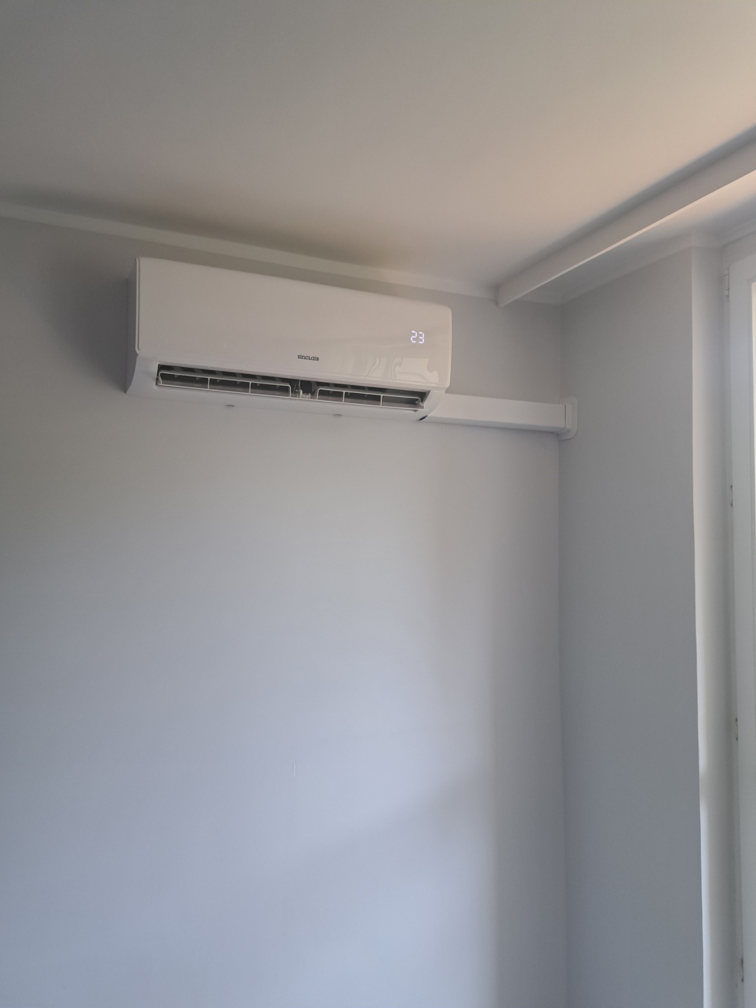 Biały klimatyzator Sinclair zamontowany na szarej ścianie w rogu pokoju, widoczny fragment okna po prawej stronie. Na urządzeniu wyświetla się temperatura 23 stopnie.