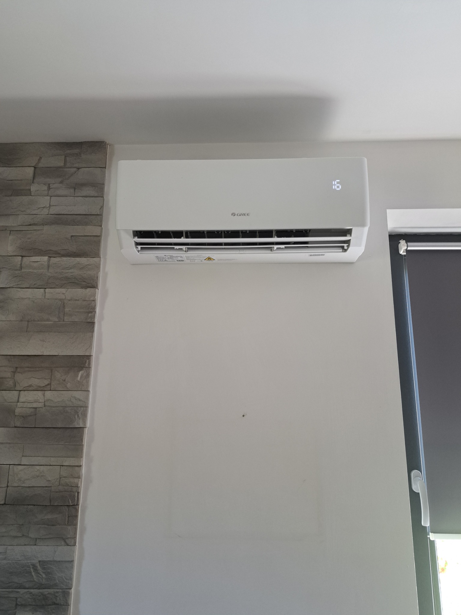 Biała klimatyzacja GREE na ścianie, obok dekoracyjna imitacja kamienia i roleta okienna. Wyświetlacz wskazuje 16 stopni. Widok z dołu.
