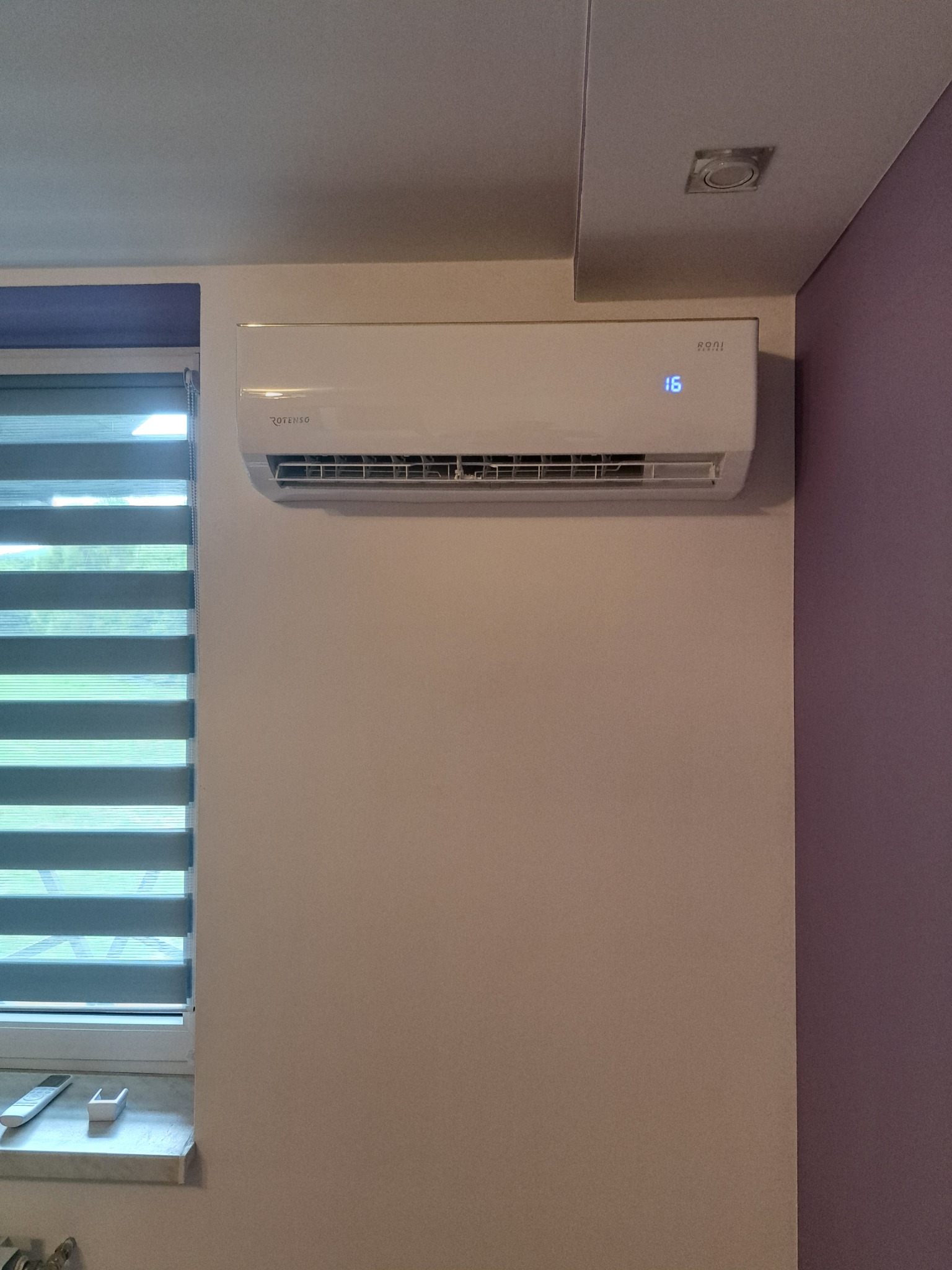 Biały klimatyzator Rotenso Roni Series zamontowany na ścianie w pokoju z roletami okiennymi i lampą sufitową. Widoczny wyświetlacz temperatury.