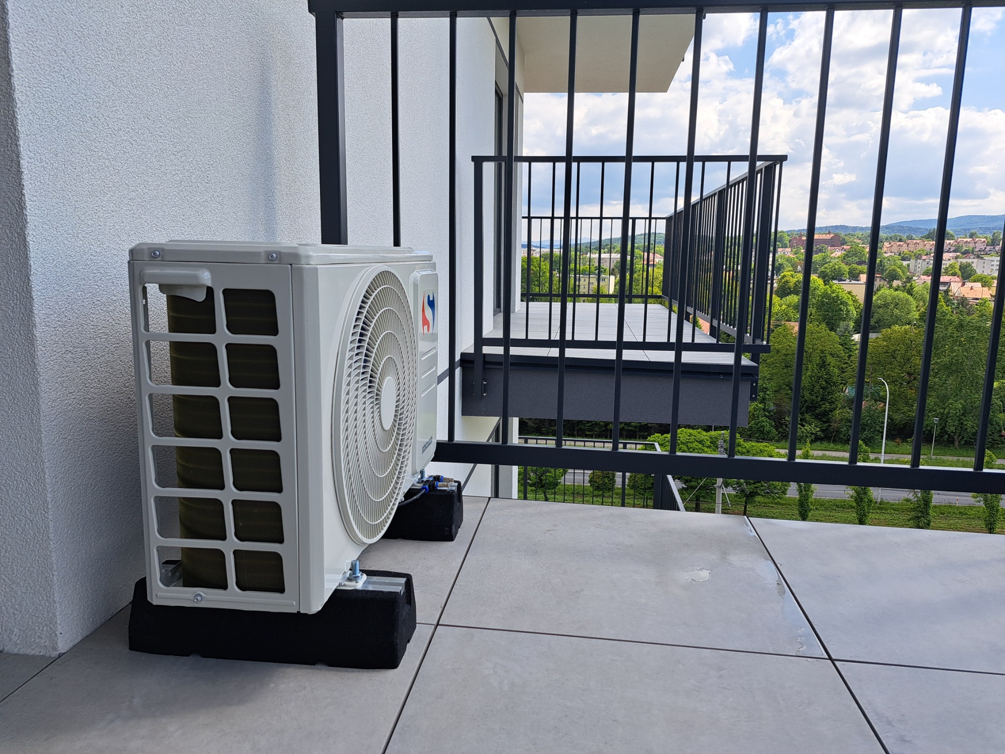 Biały klimatyzator zewnętrzny na balkonie, umieszczony na czarnych podkładkach antywibracyjnych, z widokiem na zieleń miejską przez metalową barierkę.