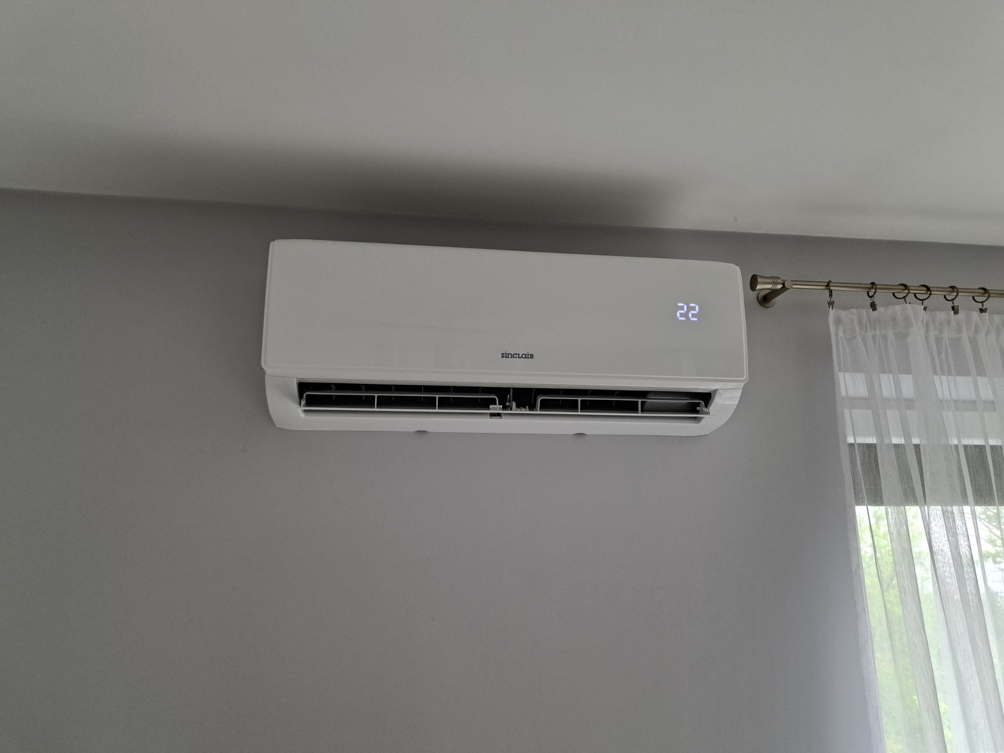 Biała klimatyzacja Sinclair na szarej ścianie, z widocznym wyświetlaczem temperatury (22 stopnie) i fragmentem firanki w oknie po prawej stronie.