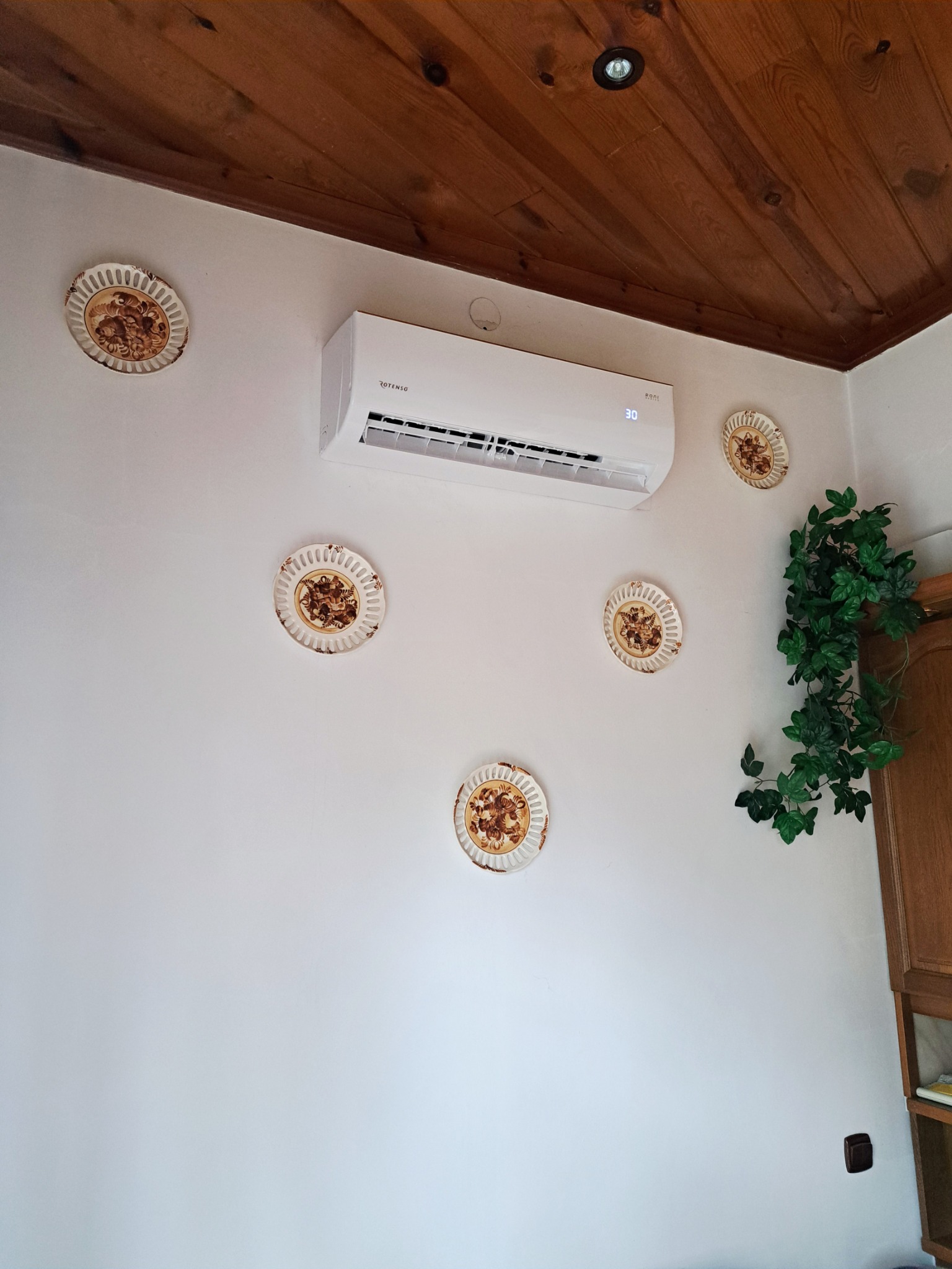 Biały klimatyzator Rotenso na ścianie z dekoracyjnymi talerzami i sztucznym bluszczem. Drewniany sufit i fragment szafki w tle.