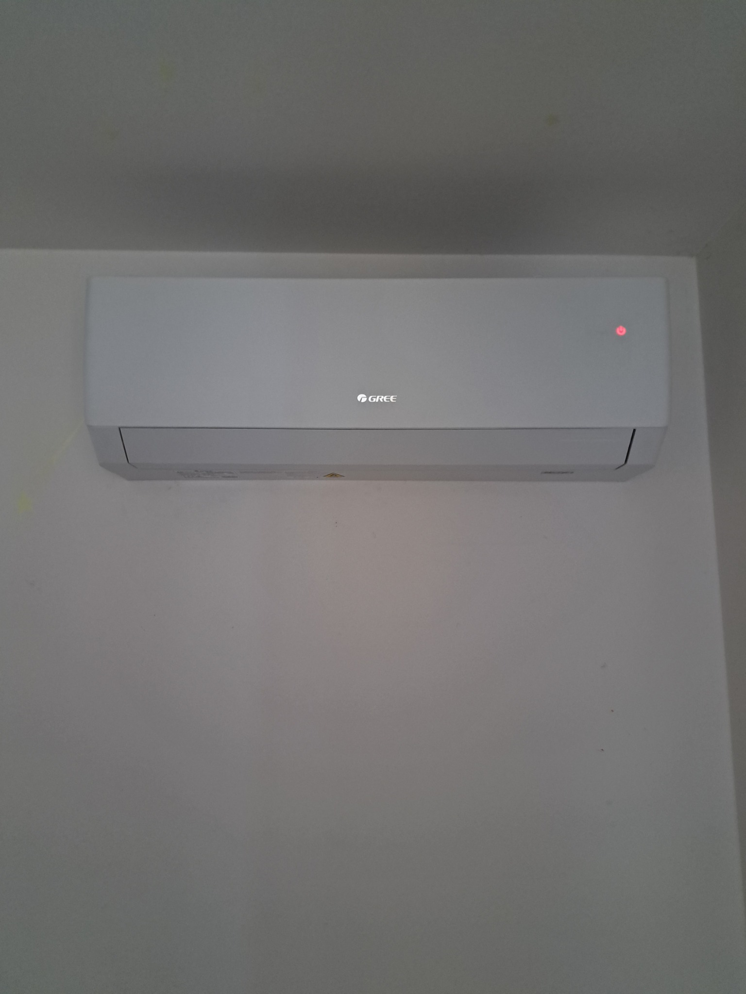 Biała klimatyzacja Gree, zamontowana na ścianie, z czerwoną diodą sygnalizacyjną. Minimalistyczny design, neutralne tło ściany.