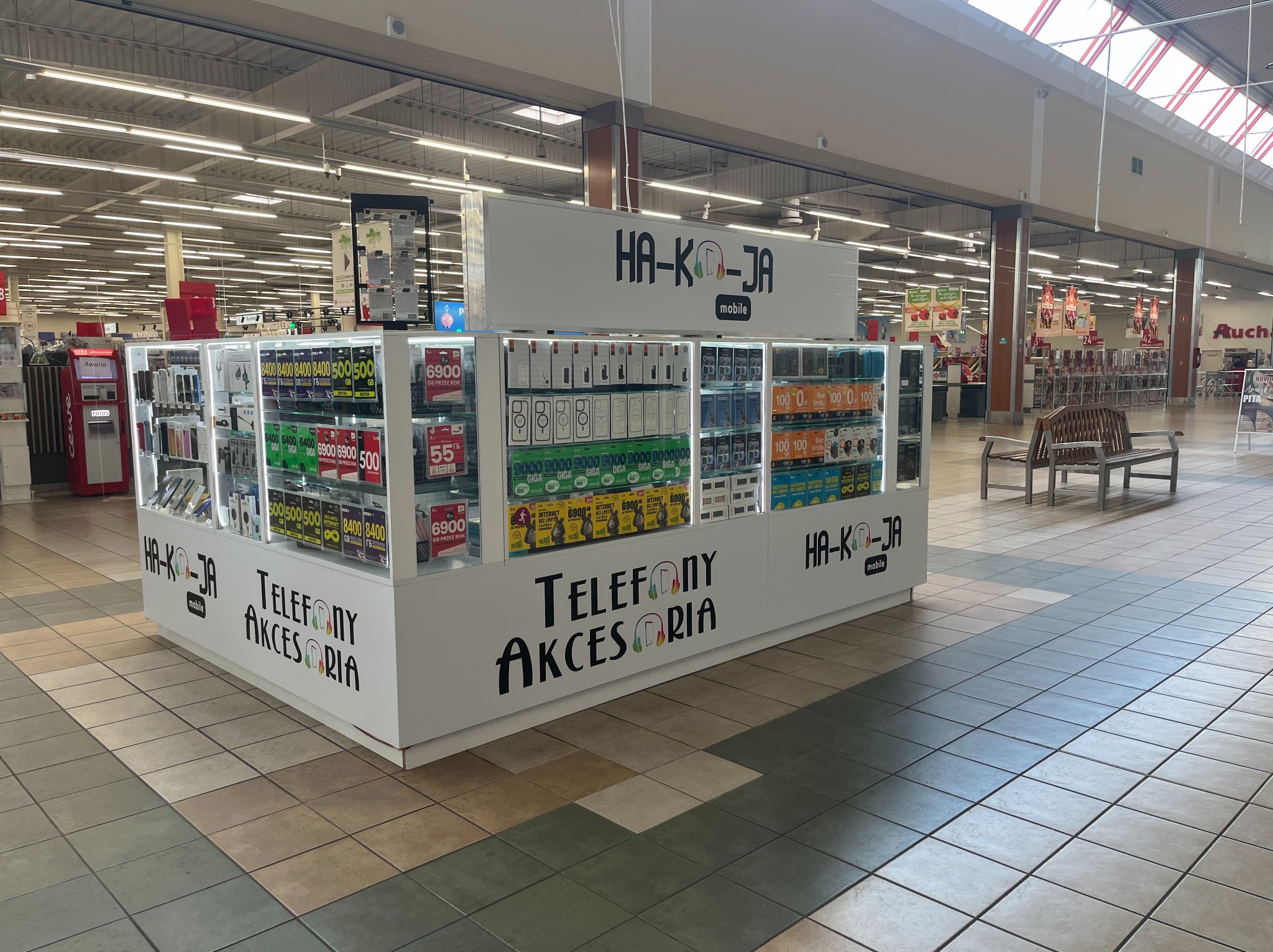 Stoisko z telefonami i akcesoriami 'HA-KO-JA mobile' w galerii handlowej. Widoczne półki z towarami i ceny. Na górze logo firmy.