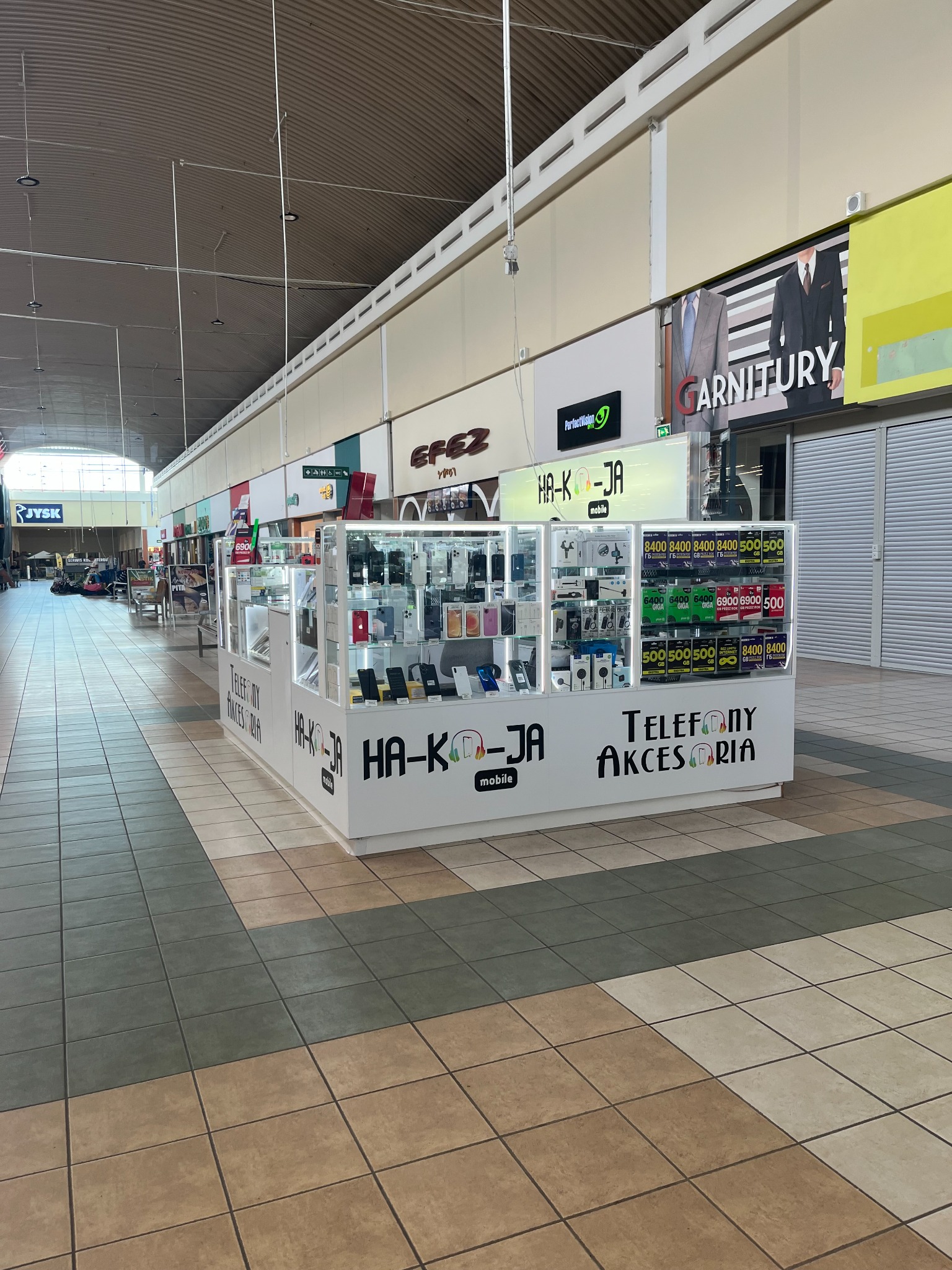 Stoisko z telefonami i akcesoriami w galerii handlowej, z widocznymi modelami telefonów i cenami w gablotach. Widok z perspektywy alei.