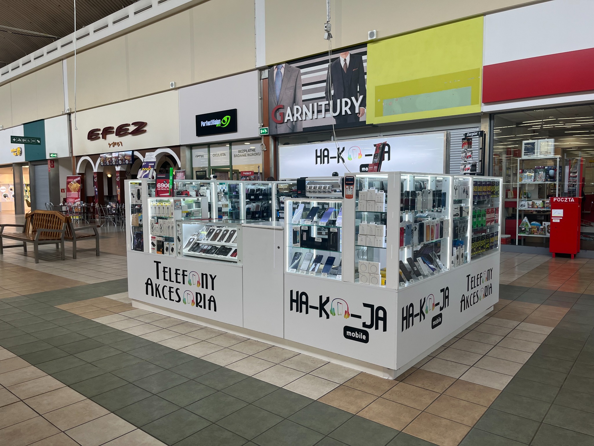 Białe stoisko z telefonami i akcesoriami w centrum handlowym, z logo 'HA-KO-JA mobile' i napisem 'Telefony Akcesoria'. Widoczne inne sklepy w tle.