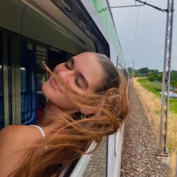 Marika Pytlewska - Uśmiechnięta, młoda kobieta z rozwianymi włosami wystawia głowę przez okno zielonego pociągu, w tle widoczne tory i krajobraz.