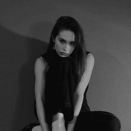 Marika Pytlewska - Monochromatyczne, studyjne ujęcie kobiety siedzącej na stołku, z nogami podciągniętymi do góry i dłońmi dotykającymi buta. Stonowana kolorystyka, minimalistyczne tło.