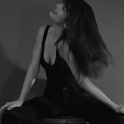 Marika Pytlewska - Monochromatyczna sesja kobieca: Kobieta w czarnym topie i spodniach siedzi na stołku, odrzucając włosy do tyłu, z uśmiechem na ustach, na tle jednolitego tła.