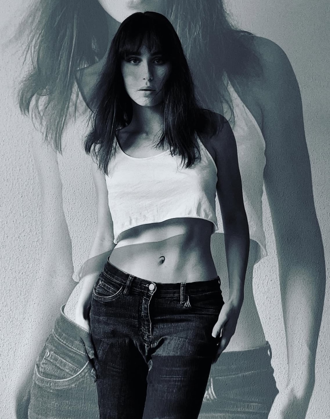 Czarno-białe zdjęcie kobiety w topie i dżinsach, stojącej na tle ściany z widocznym cieniem, tworzącym efekt podwójnej ekspozycji.