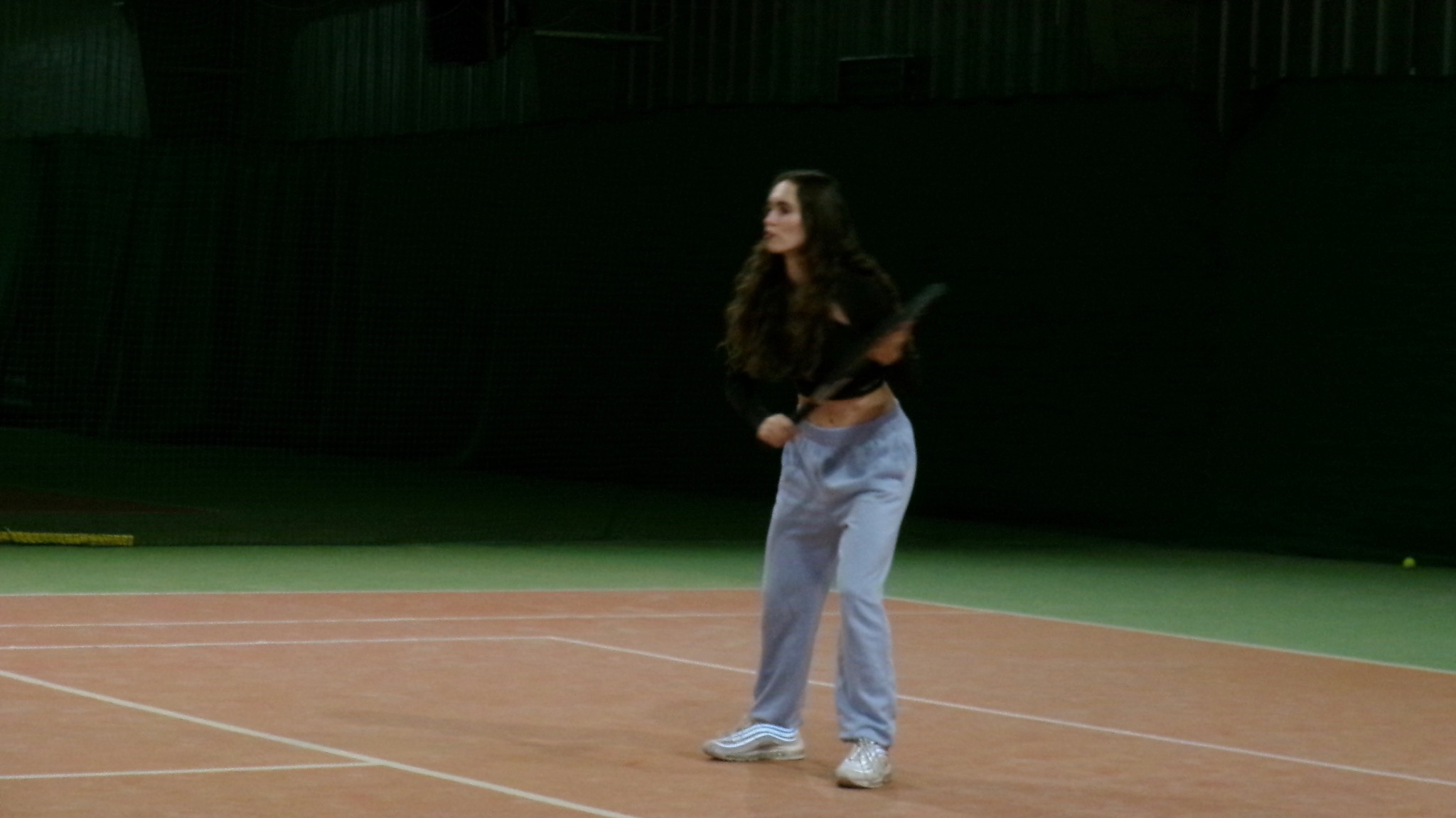 Kobieta z długimi włosami, ubrana w sportowy strój, w trakcie serwowania na korcie tenisowym. Ujęcie z boku, widoczna rakieta i piłka w tle.