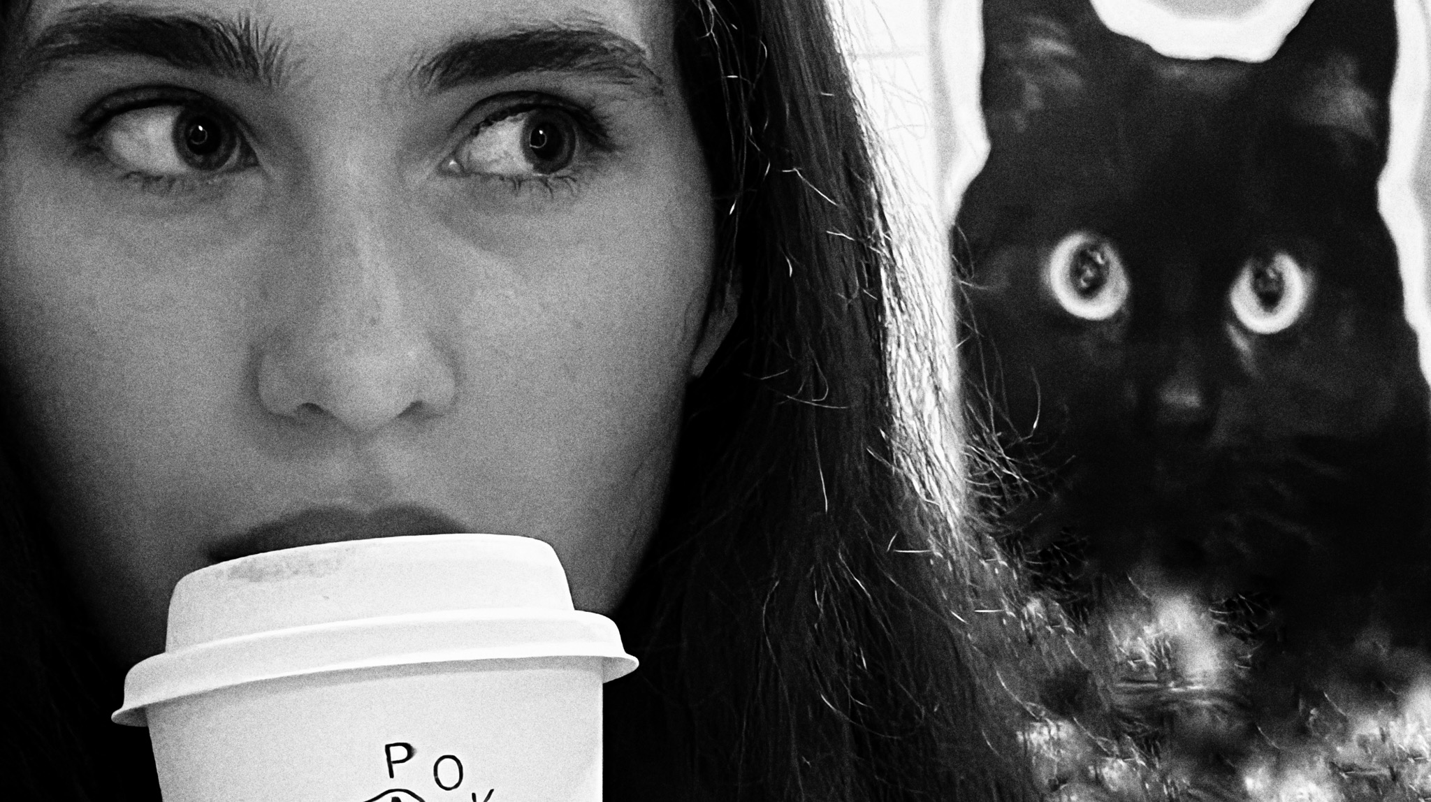 Czarno-białe zbliżenie kobiety z kawą, spoglądającej w bok, z sylwetką kota w tle. Nietypowe ujęcie, kontrastowe oświetlenie.